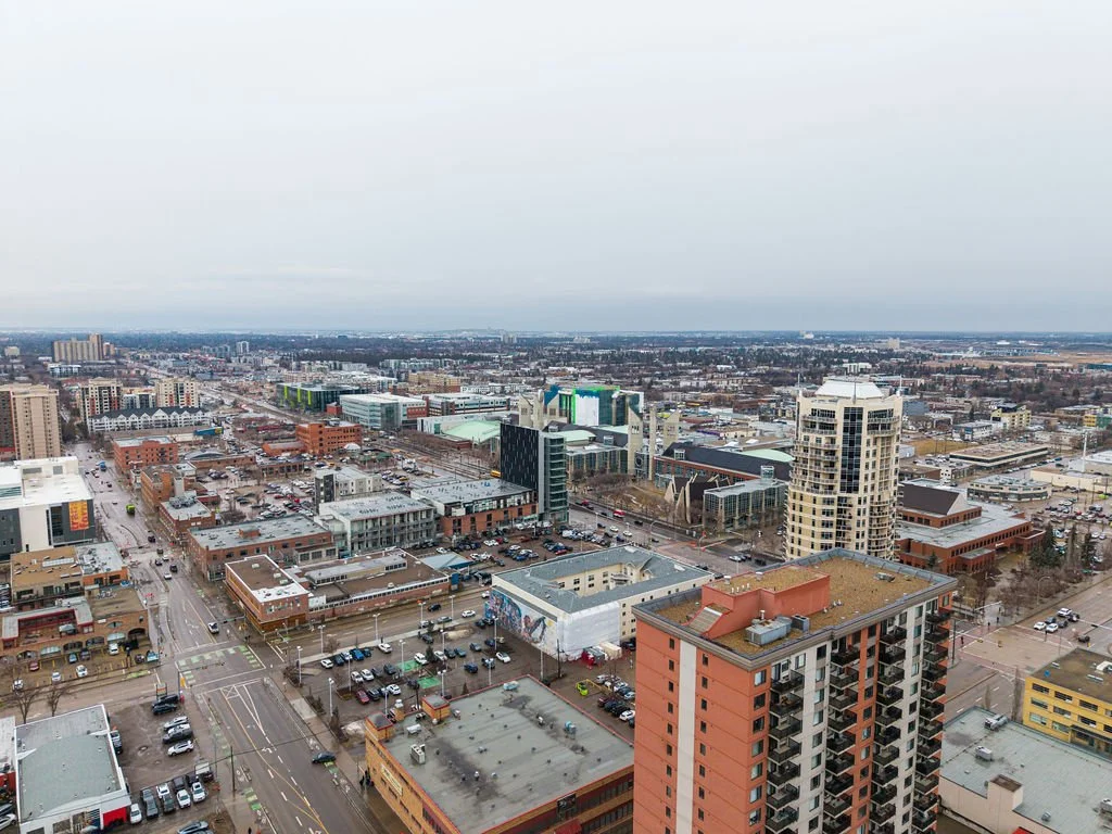Aerial view of a cityscape showing various commercial and residential buildings, streets, and parking lots under an overcast sky.