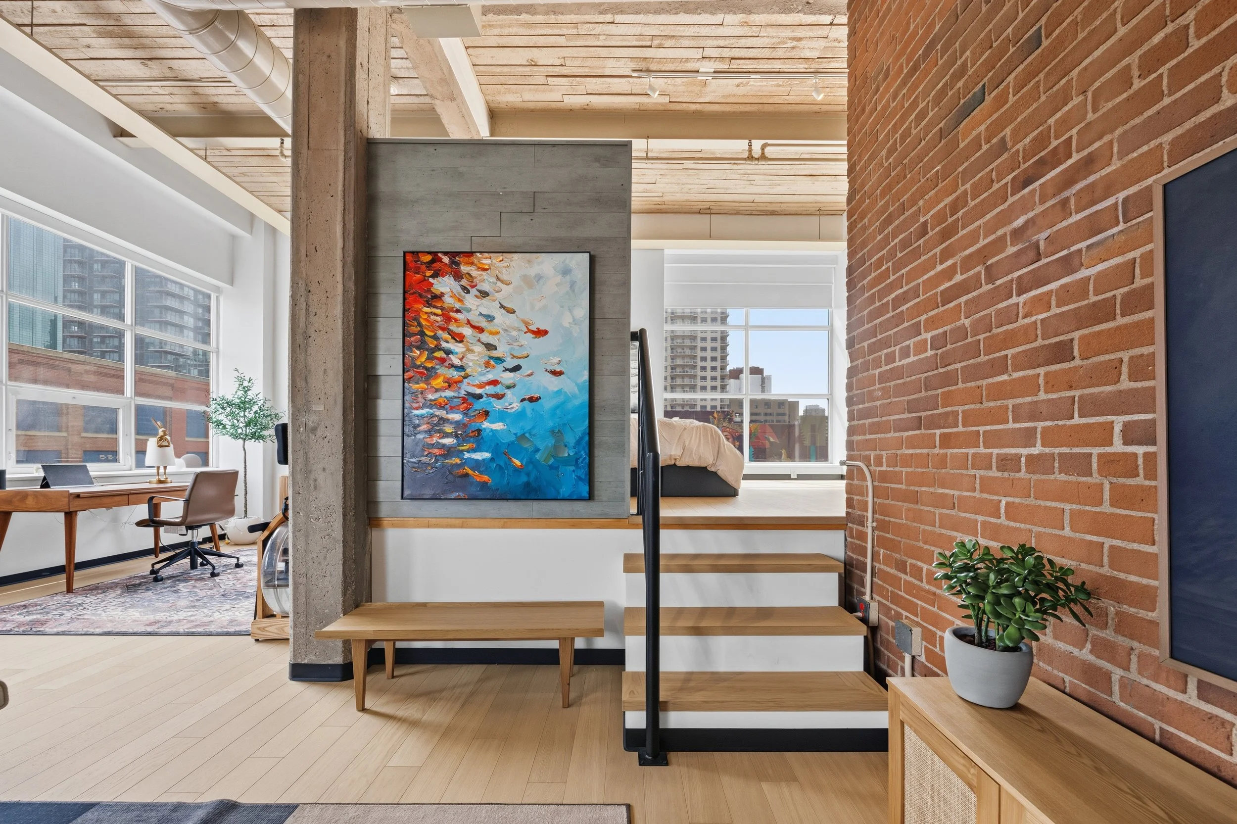 Interior of a modern urban apartment with exposed brick and concrete walls, large windows, artwork, plants, and wooden stairs.