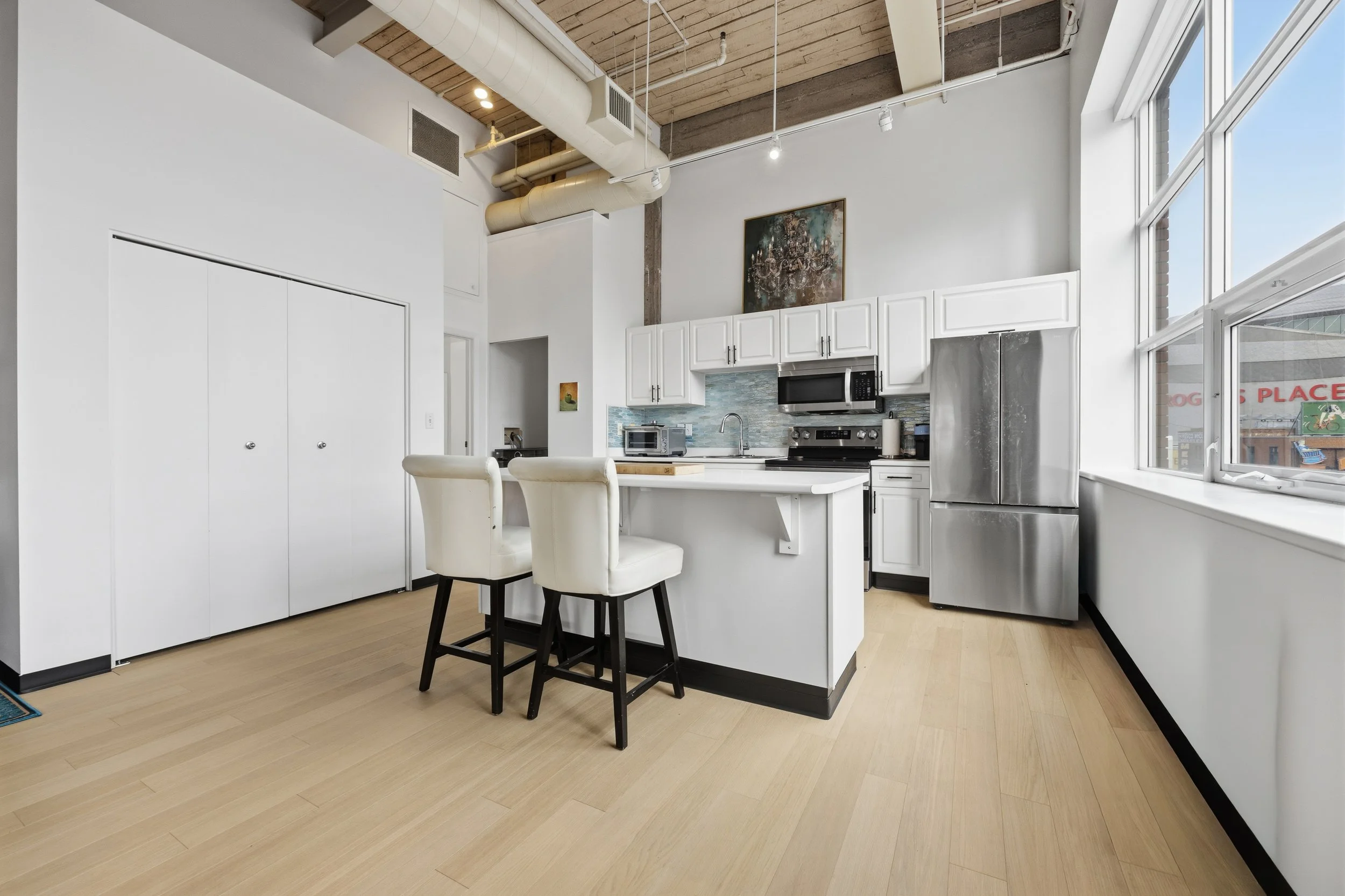 Modern kitchen with white cabinets, stainless steel appliances, a small dining table with two white chairs, large windows, and exposed ceiling pipes and wooden beams.