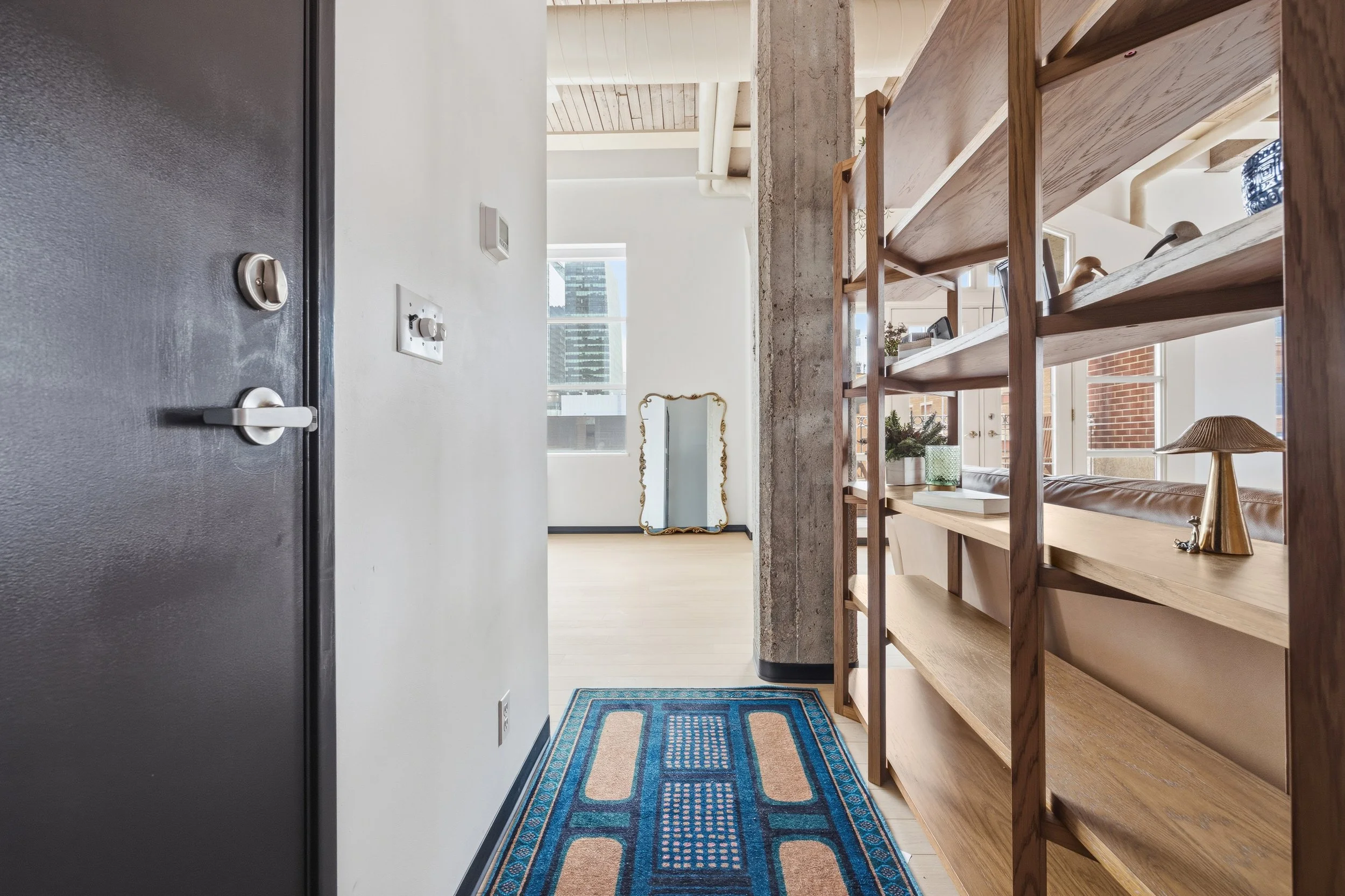 View of a bright, modern apartment hallway with large windows, wood shelving, a decorative mirror, and a rug.
