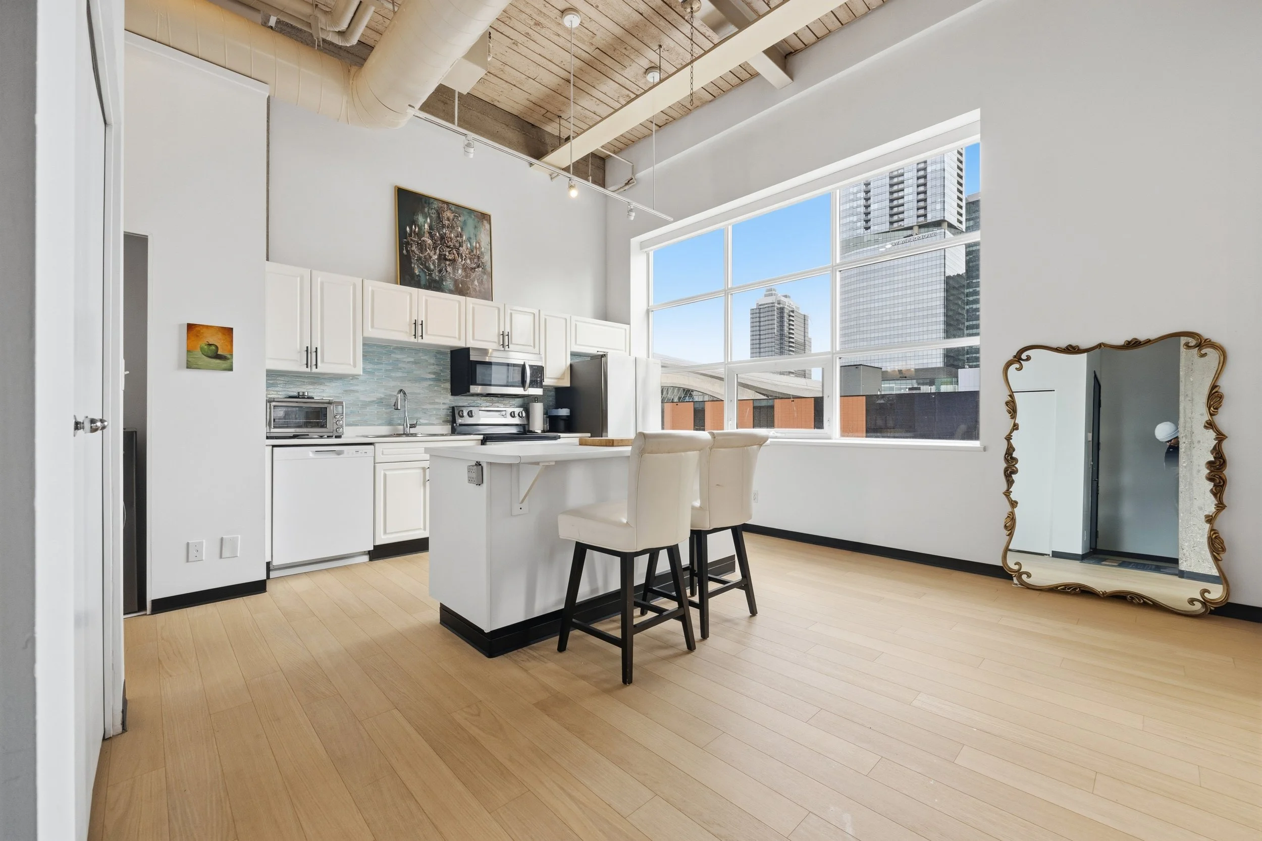 Interior view of a modern apartment or condo with high ceilings, large windows, and a cityscape view. The space includes a kitchen with white cabinets, a small island with two chairs, and artwork on the wall. There is a large, ornate mirror leaning a