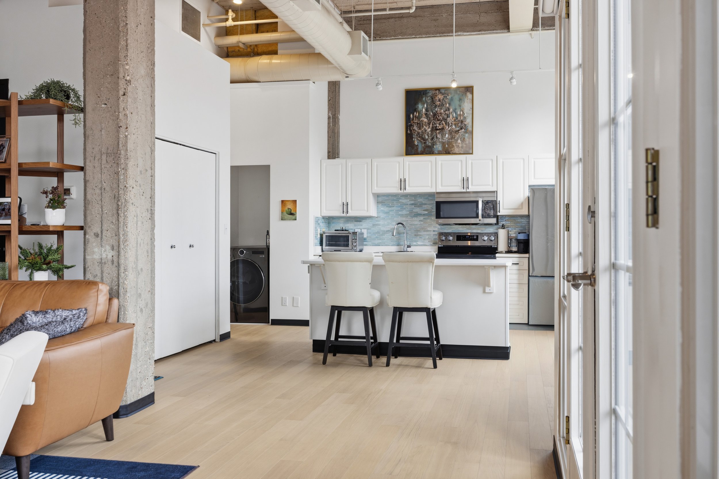 Modern kitchen with white cabinets, stainless steel appliances, and a painting on the wall. An island with two bar stools is in the center. Part of a living room with a brown leather chair and shelving with framed photos is visible in the foreground.