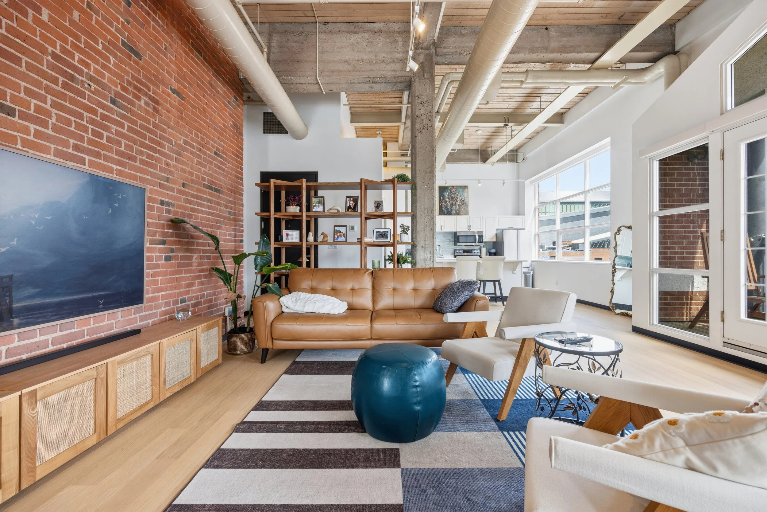 Bright living room with exposed brick wall, wooden ceiling, large windows, and modern furniture including a tan leather sofa, white chairs, a blue ottoman, and a striped rug. There is a wooden shelving unit with framed photos and decorative items, an