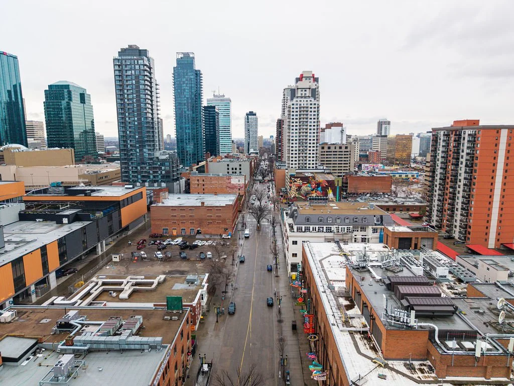 City skyline with tall modern glass buildings, a street with parked cars, and smaller brick buildings in an urban area on a cloudy day.