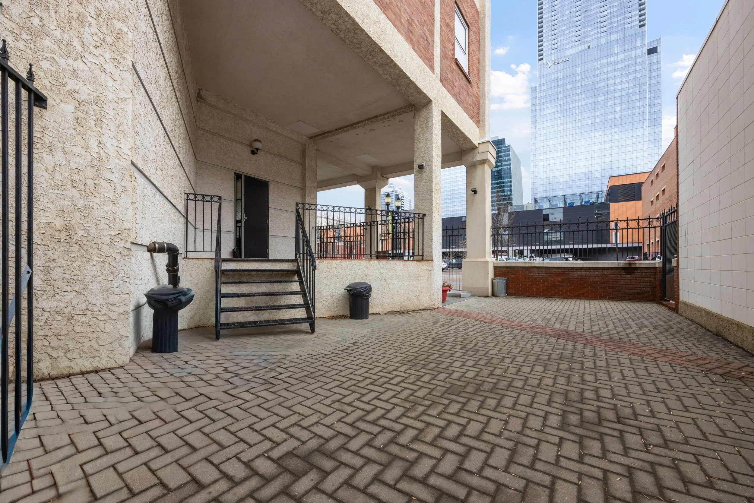 Urban outdoor scene featuring a brick and stucco building with a second story balcony, black metal staircase, trash cans, and a paved courtyard surrounded by high-rise glass buildings on a partly cloudy day.