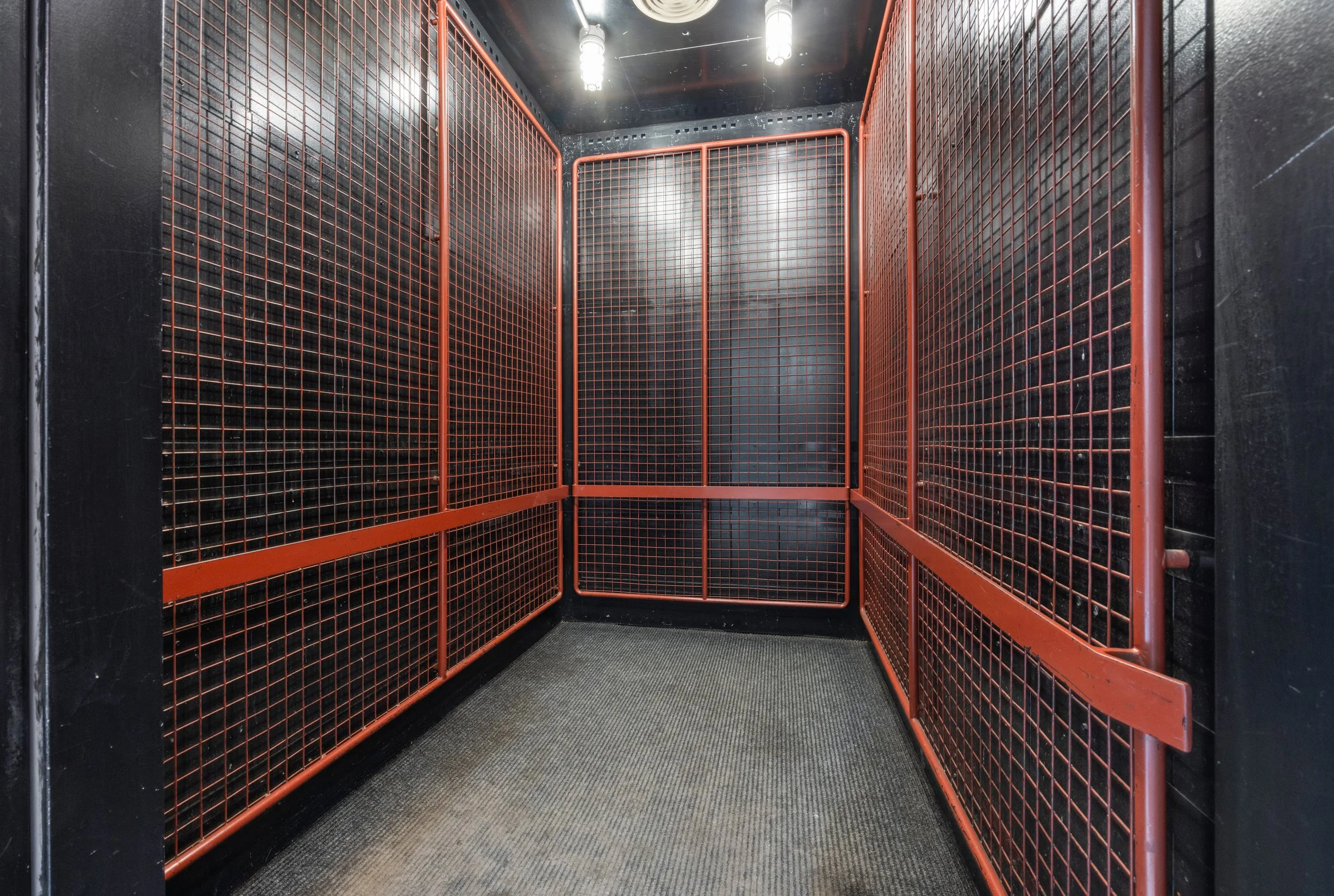 An interior view of a small elevator with orange metal mesh walls, black walls, a black ceiling, and patterned carpet flooring. Fluorescent lights are on the ceiling.
