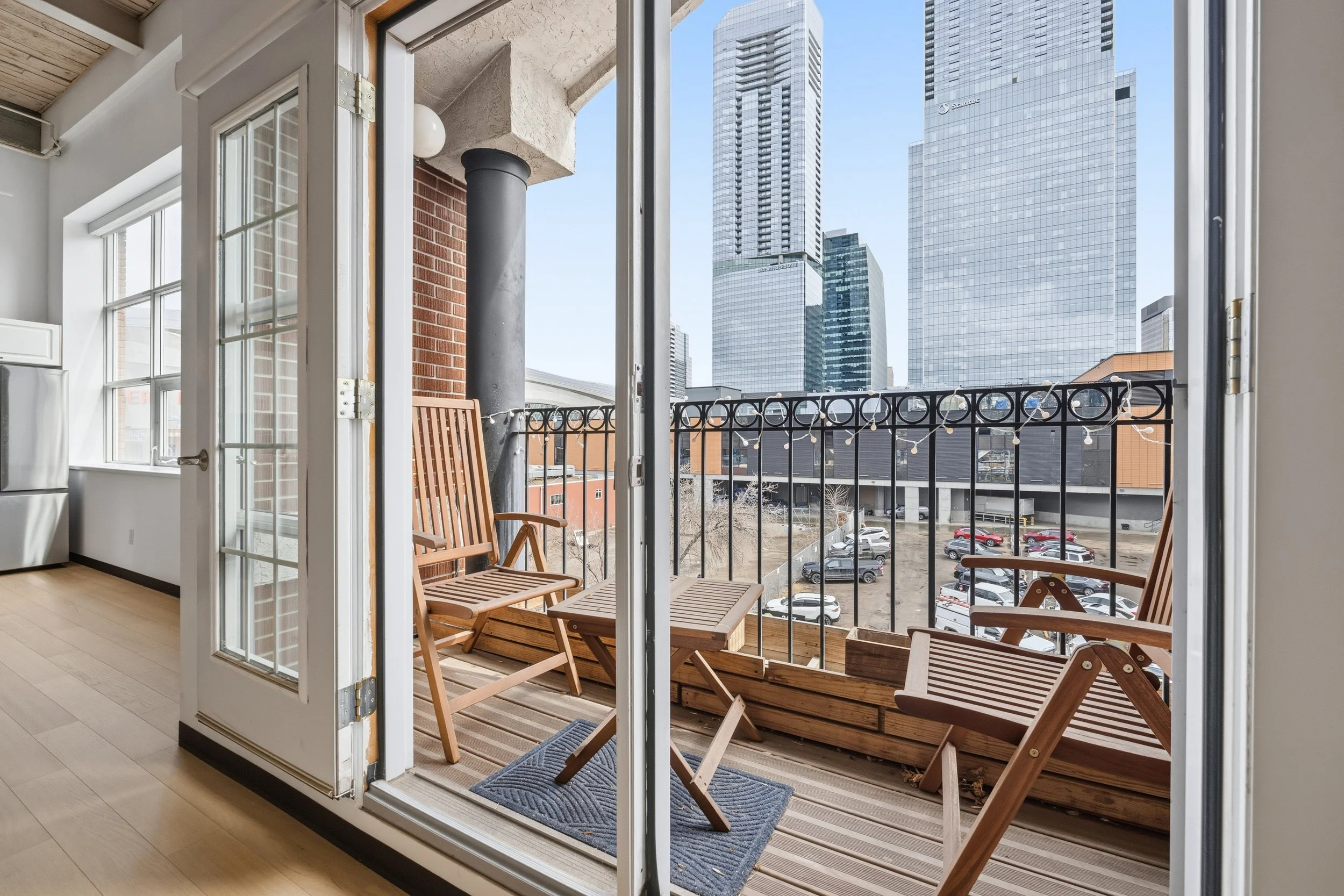 A small balcony with wooden chairs and a plant box, overlooking a parking lot and modern skyscrapers.