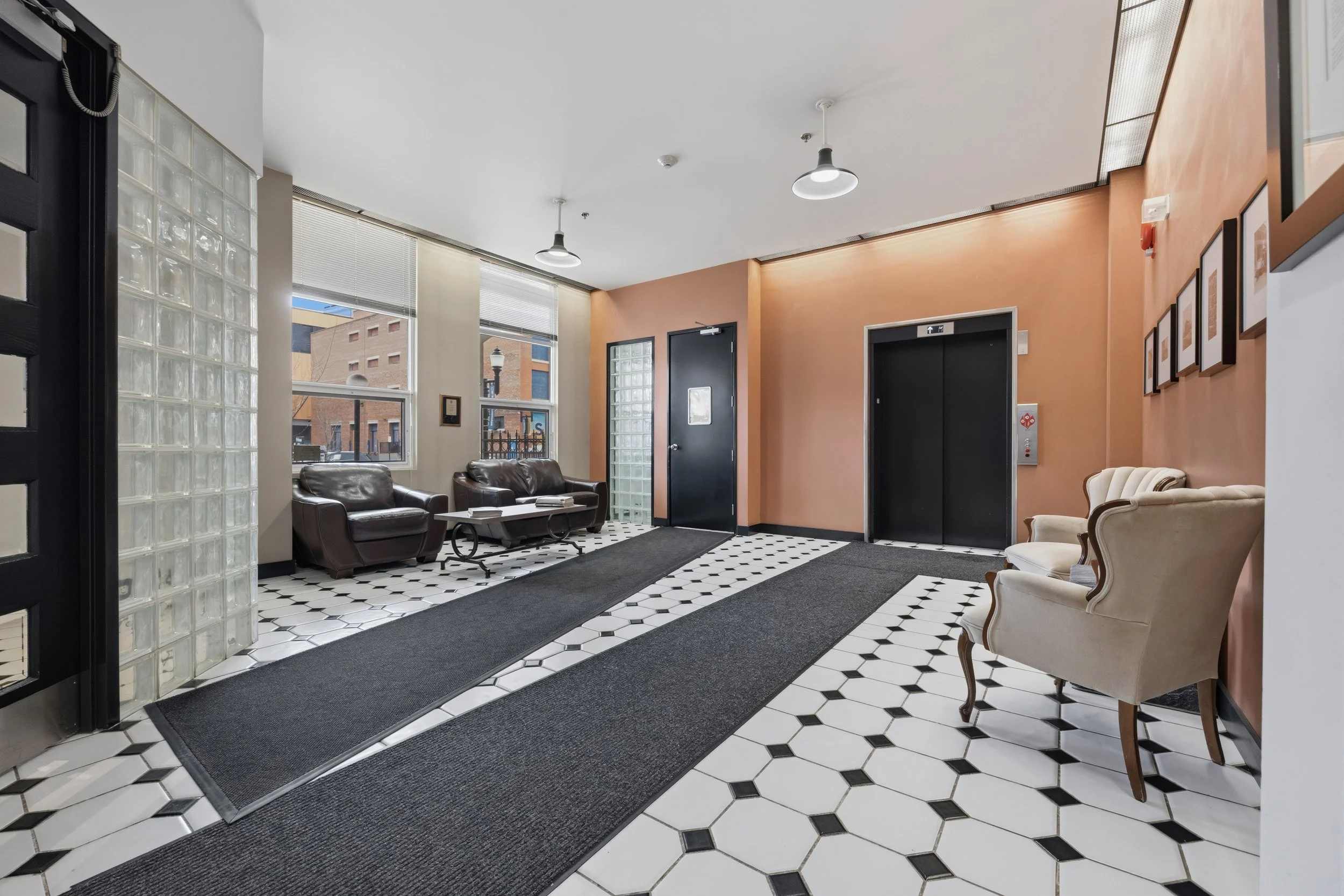 Lobby with black and white hexagonal tile flooring, black elevator doors, beige walls, chairs, and large windows.