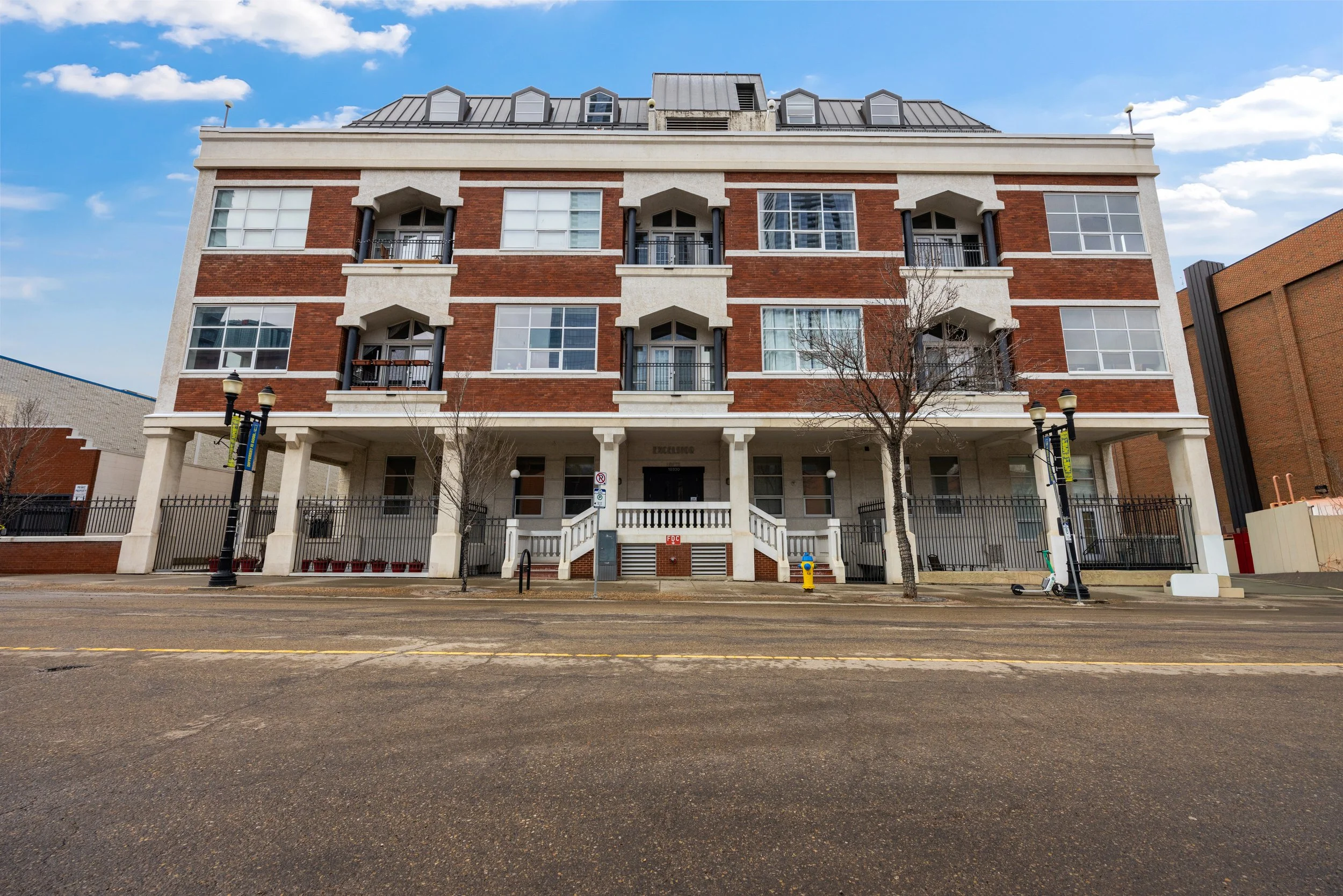 A multi-story brick building with large windows and small balconies, surrounded by a black fence and street lamps, with a cloudy blue sky above.