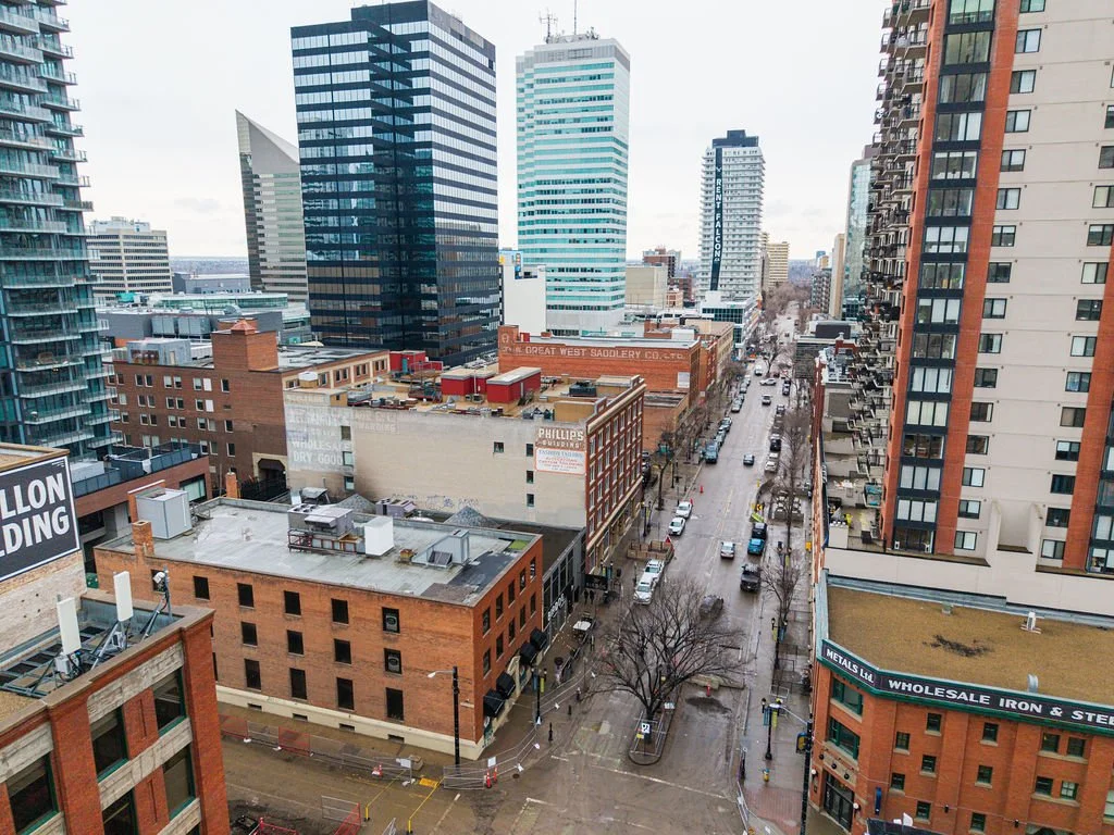 A city street viewed from above, lined with tall buildings, with some trees along the sidewalk and cars parked or driving on the street. Tall office buildings and residential high-rises are visible in the background.