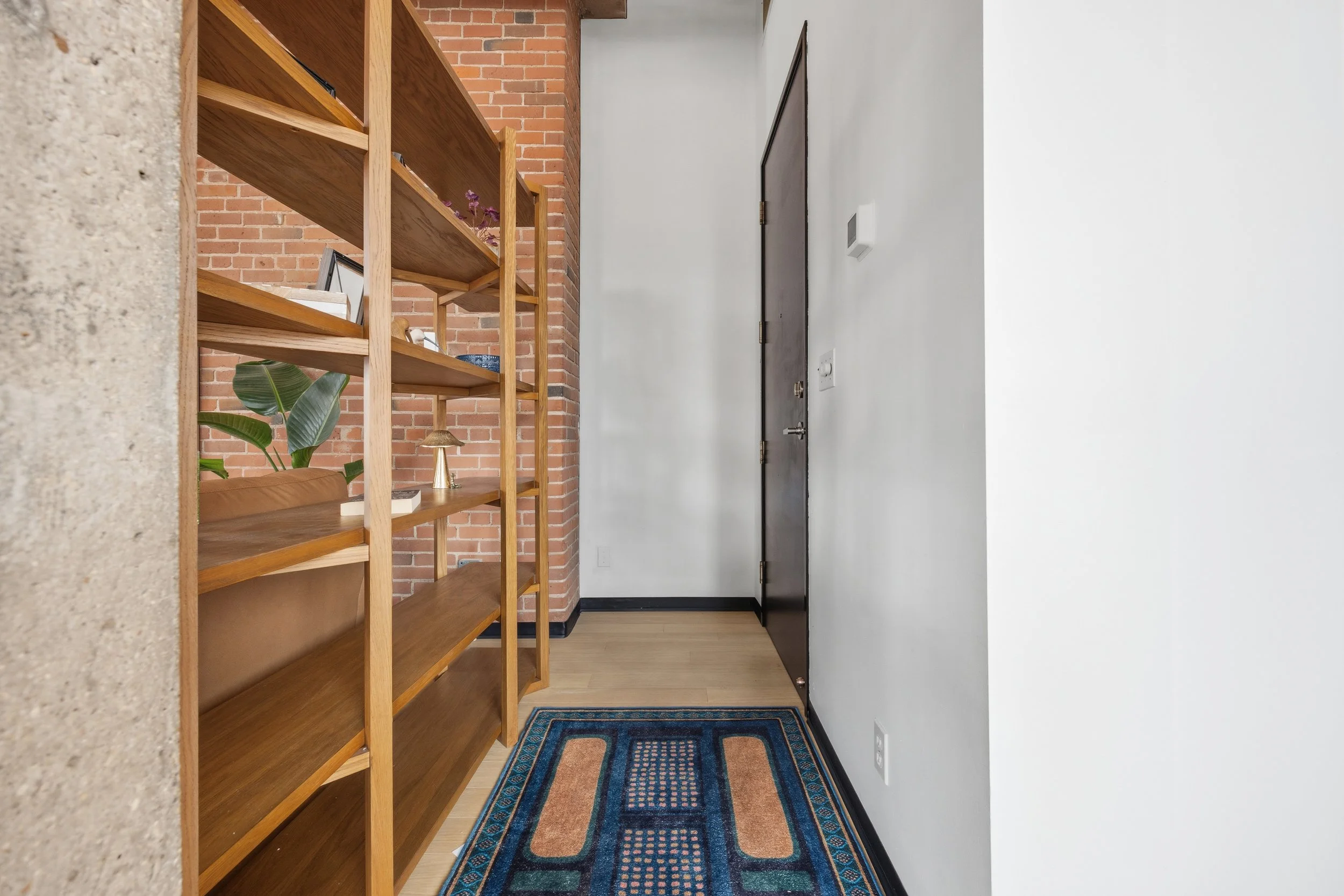 A narrow hallway with a wooden bookshelf on the left, brick wall behind it, white wall on the right, and a dark door at the far end. There is a colorful patterned rug on the light wooden floor near the entrance.