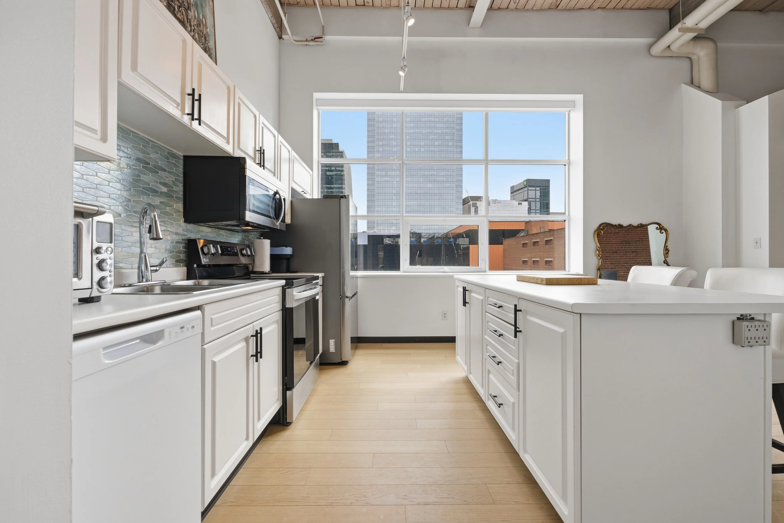 Modern kitchen with white cabinets, black handles, stainless steel appliances, and a large window showcasing city buildings.