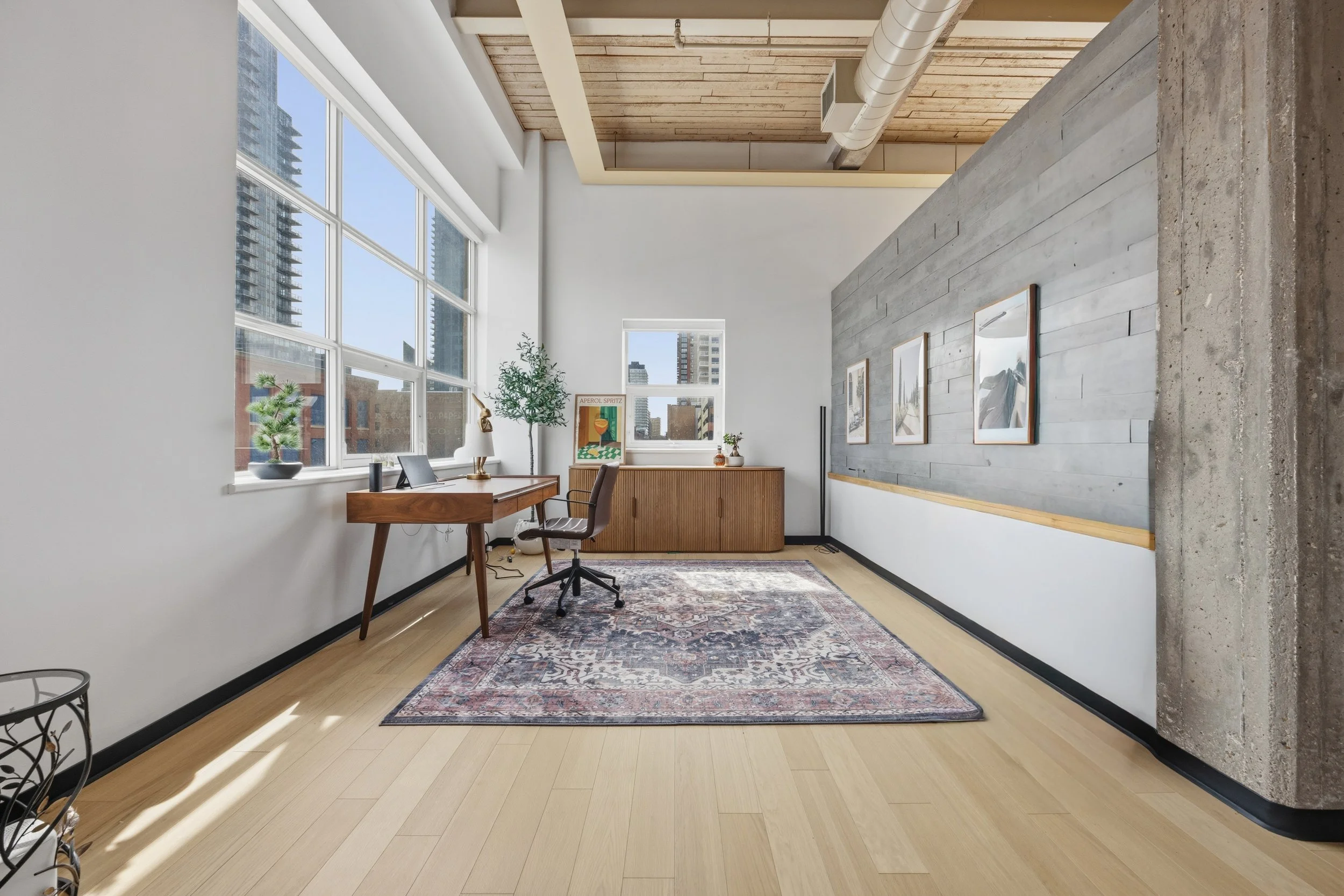 A modern office space with large windows allowing natural light, a wooden desk with a computer and lamp, a chair, potted plants, framed artwork on a textured gray wall, a decorative rug, and an exposed wooden ceiling with industrial ductwork.