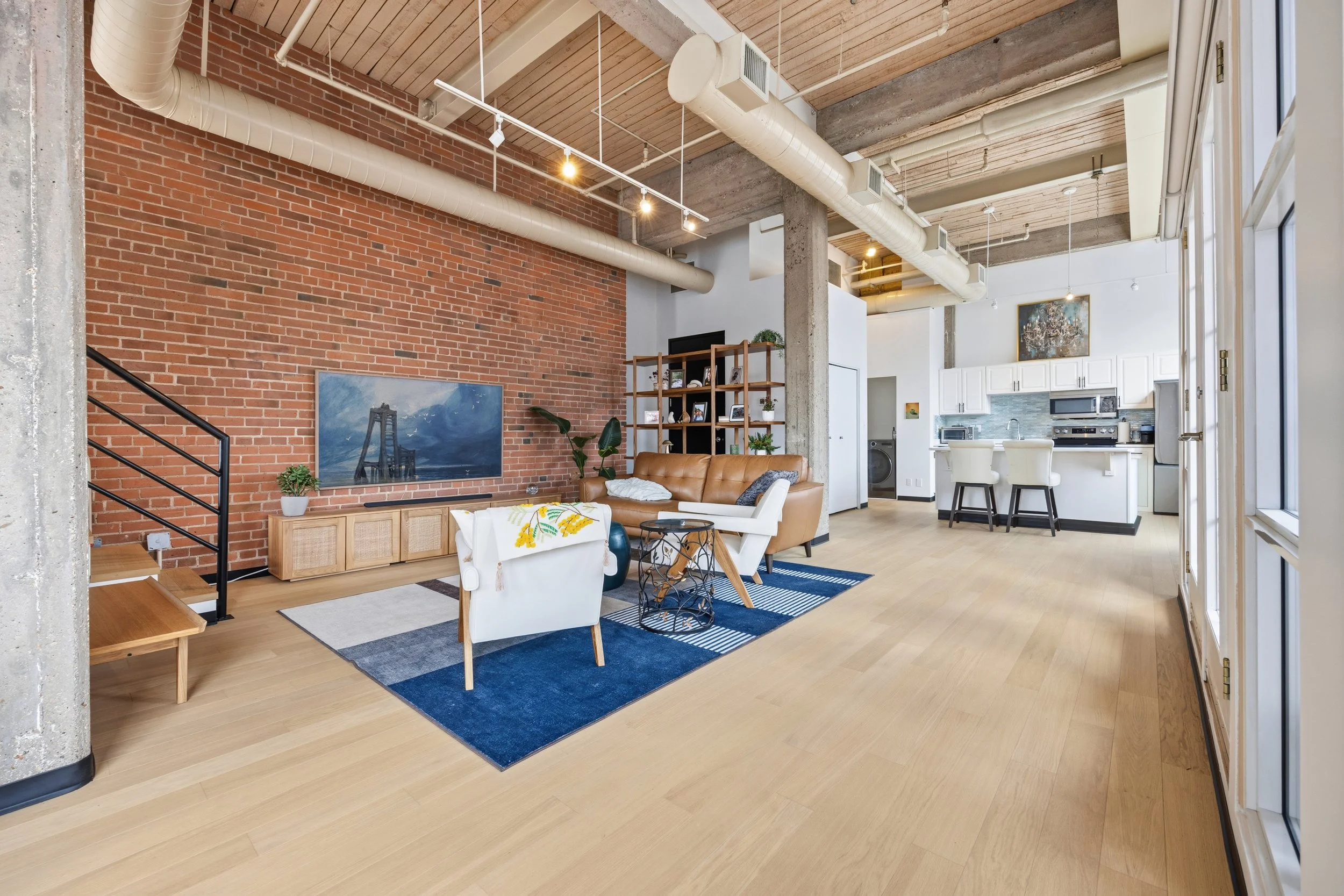 Open-concept living space with brick wall, wood accents, modern kitchen, and dining area, featuring a leather sofa, white chairs, and decorative shelving.