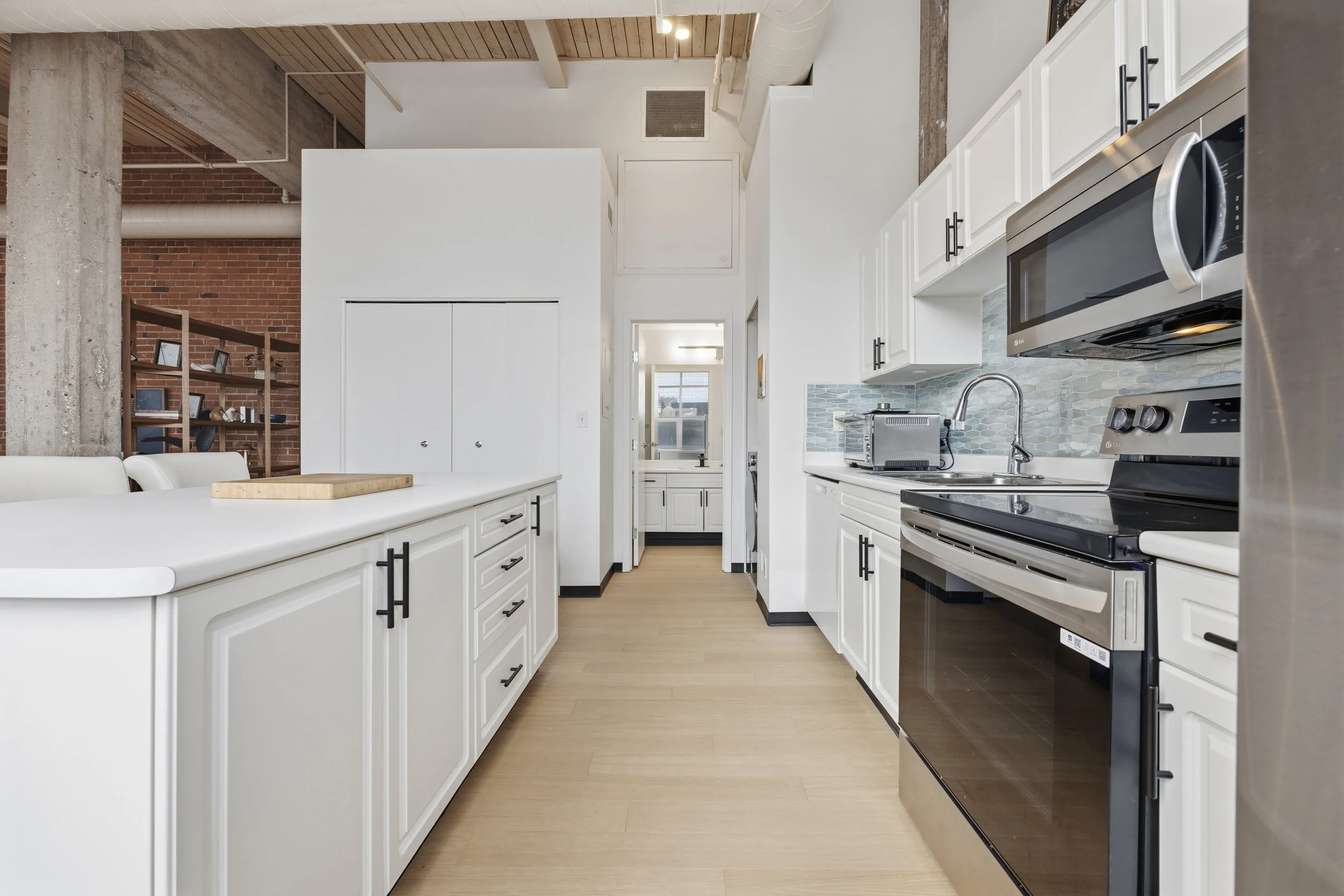 Modern kitchen with white cabinets, stainless steel appliances, tiled backsplash, and light wood flooring, with exposed brick and concrete elements in the background.