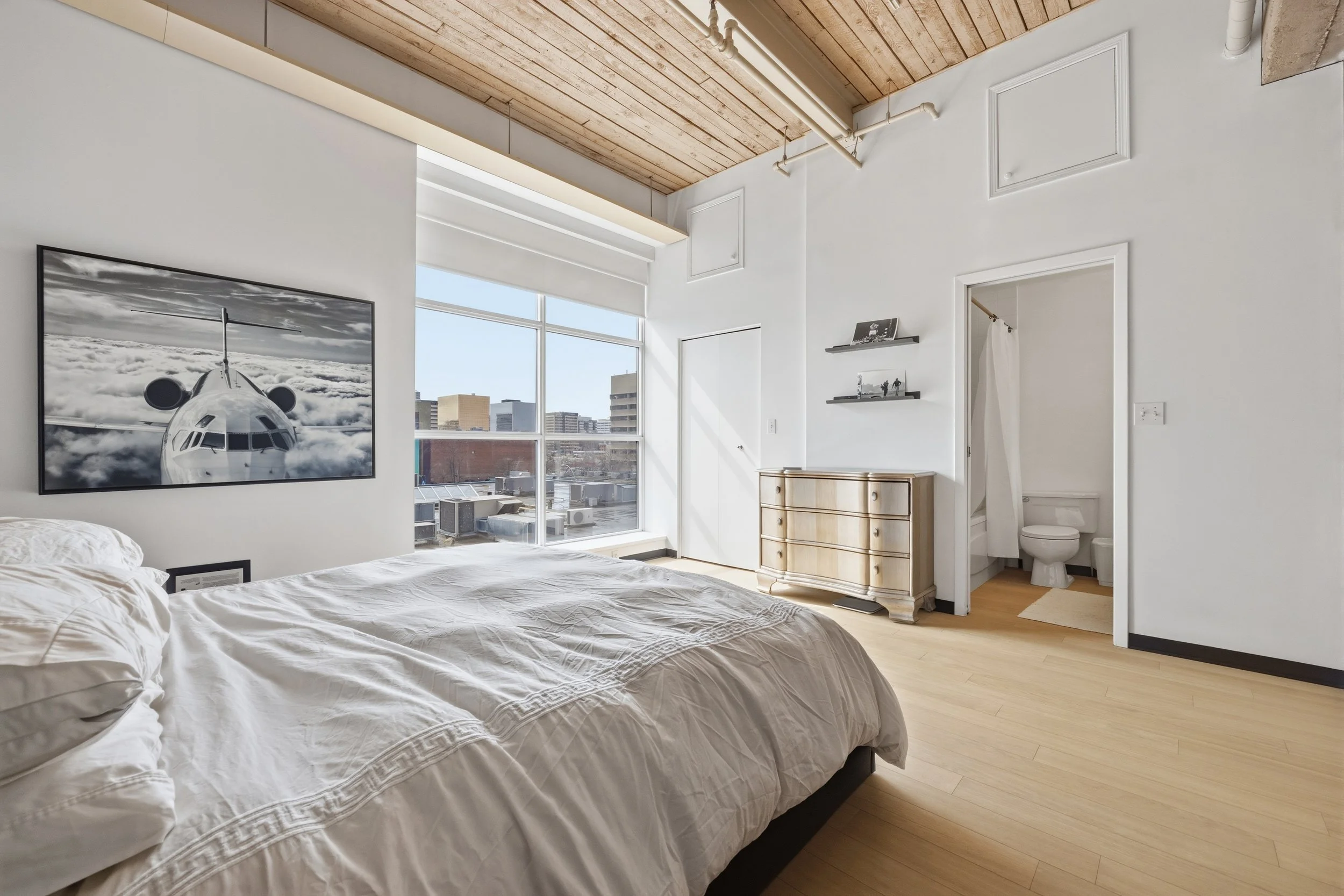A modern bedroom with a large window showing a cityscape, a bed with white bedding, a wooden dresser, and a small bathroom with a toilet visible through an open door. Art of a jet airplane is hung on the wall.
