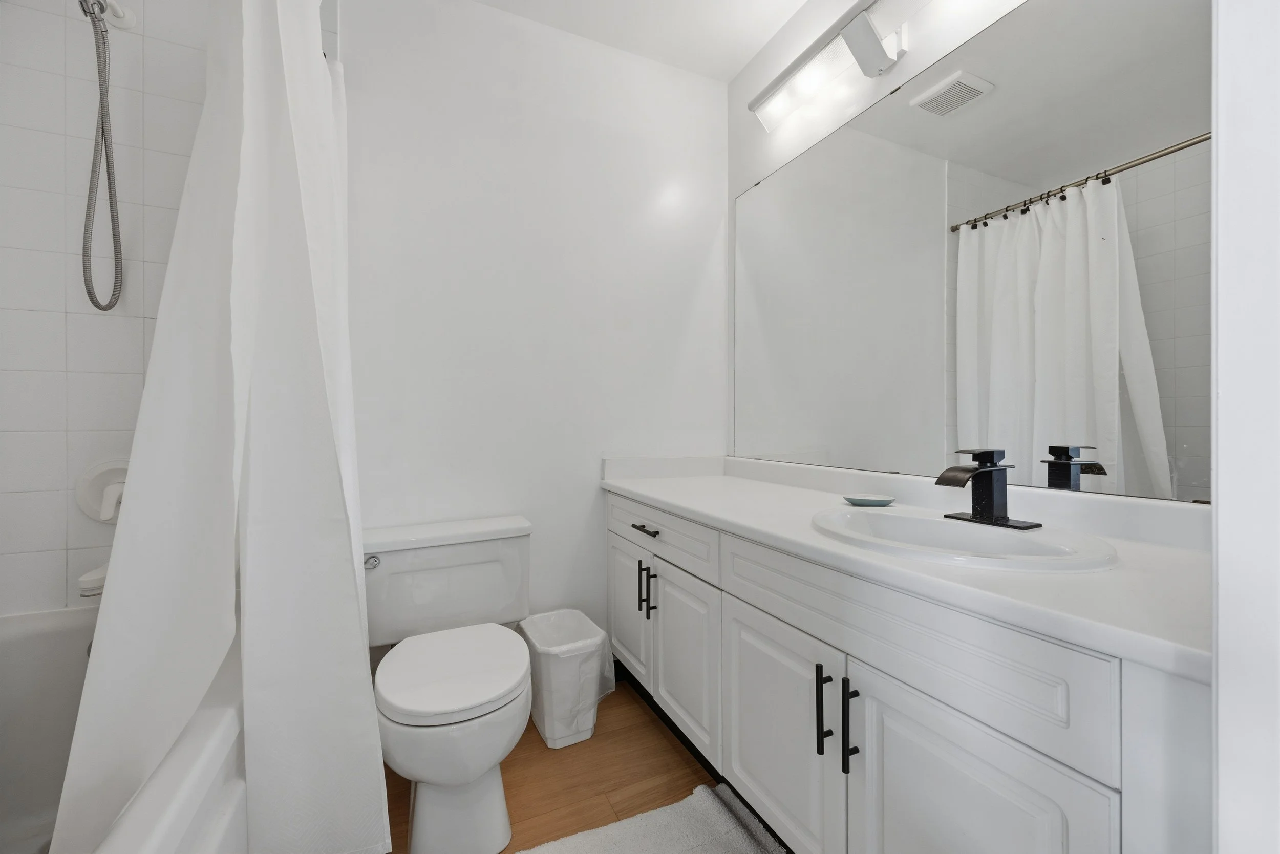 A small, white bathroom with a shower on the left, a toilet below a mirror, and a white vanity with black handles on the right. The shower has a white curtain and a handheld showerhead. The bathroom has wood flooring and a white towel on the floor.