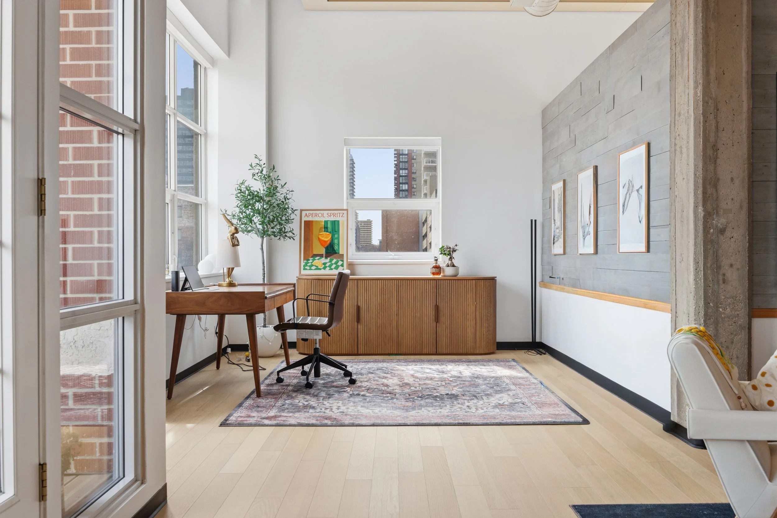 Interior of a bright living space with large windows, a wooden desk with a chair, and decorative art on the wall. There is a potted plant and framed artwork on a side table near the window.