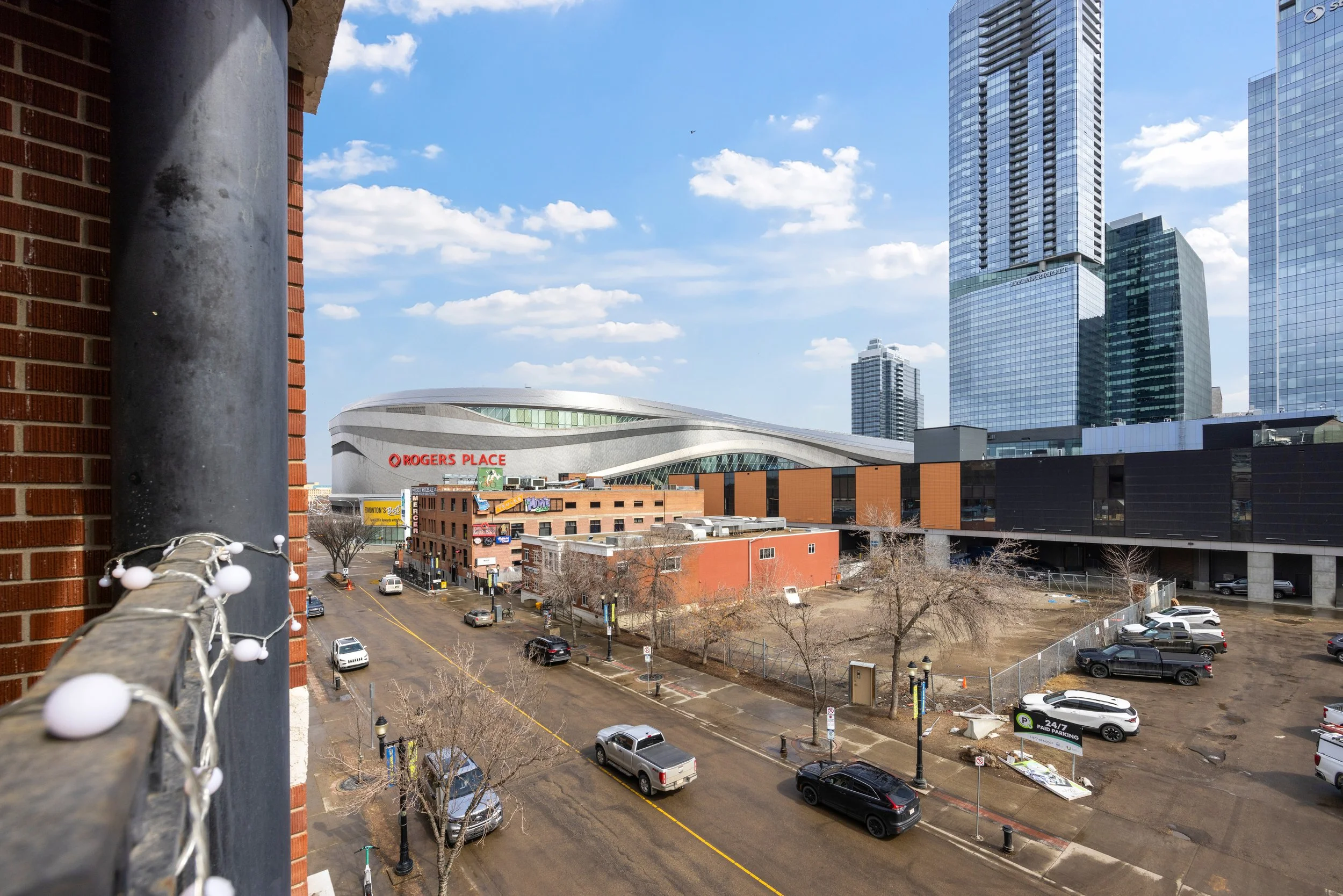 View of a cityscape with modern tall buildings, a stadium labeled Rovgers Place, parking lots, cars, and trees, taken from a high-rise balcony with a brick wall and string lights in the foreground.