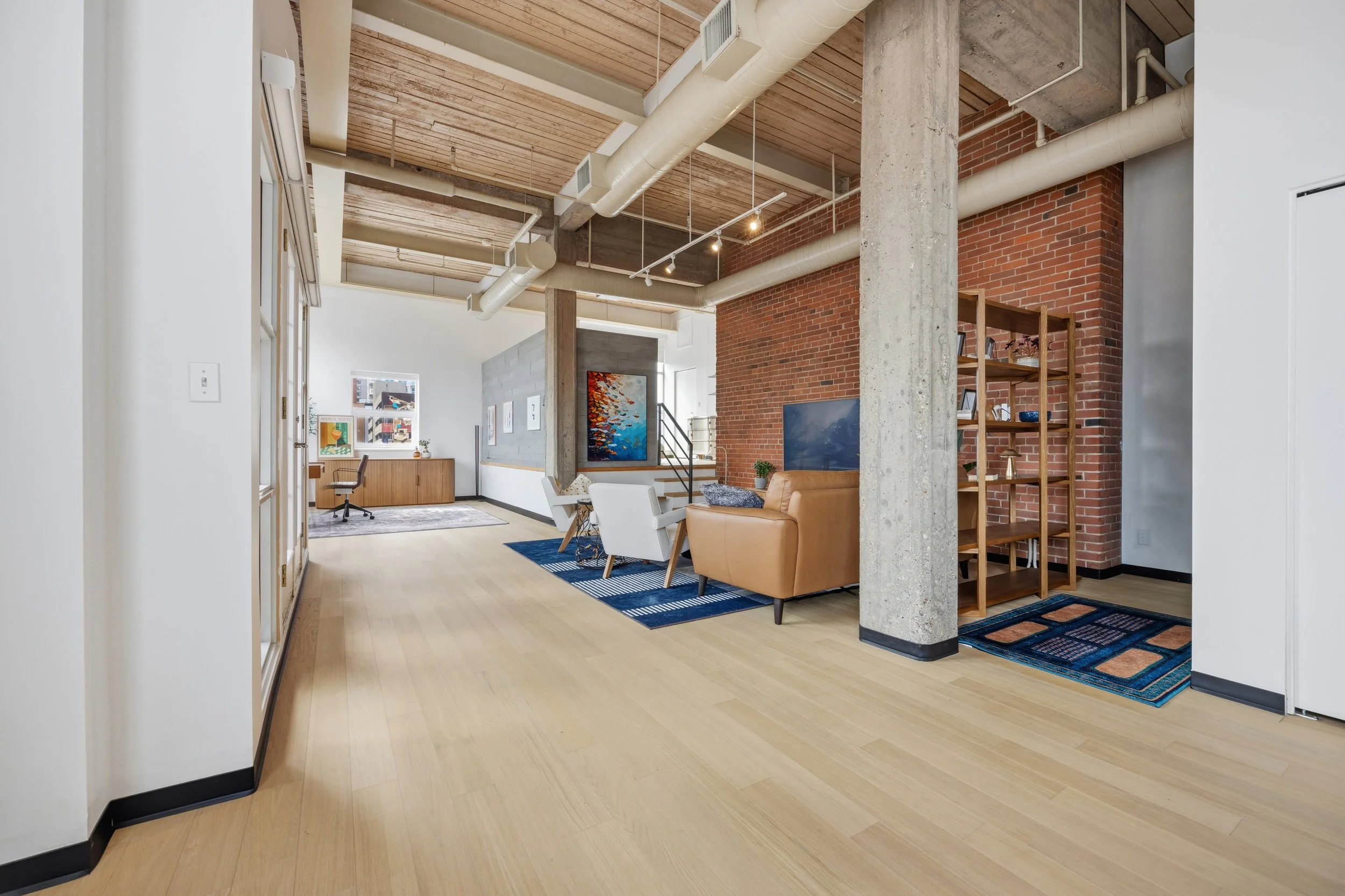Open-plan modern loft apartment with exposed brick and concrete walls, wooden ceiling, and minimalist furniture including leather couch and white chairs.