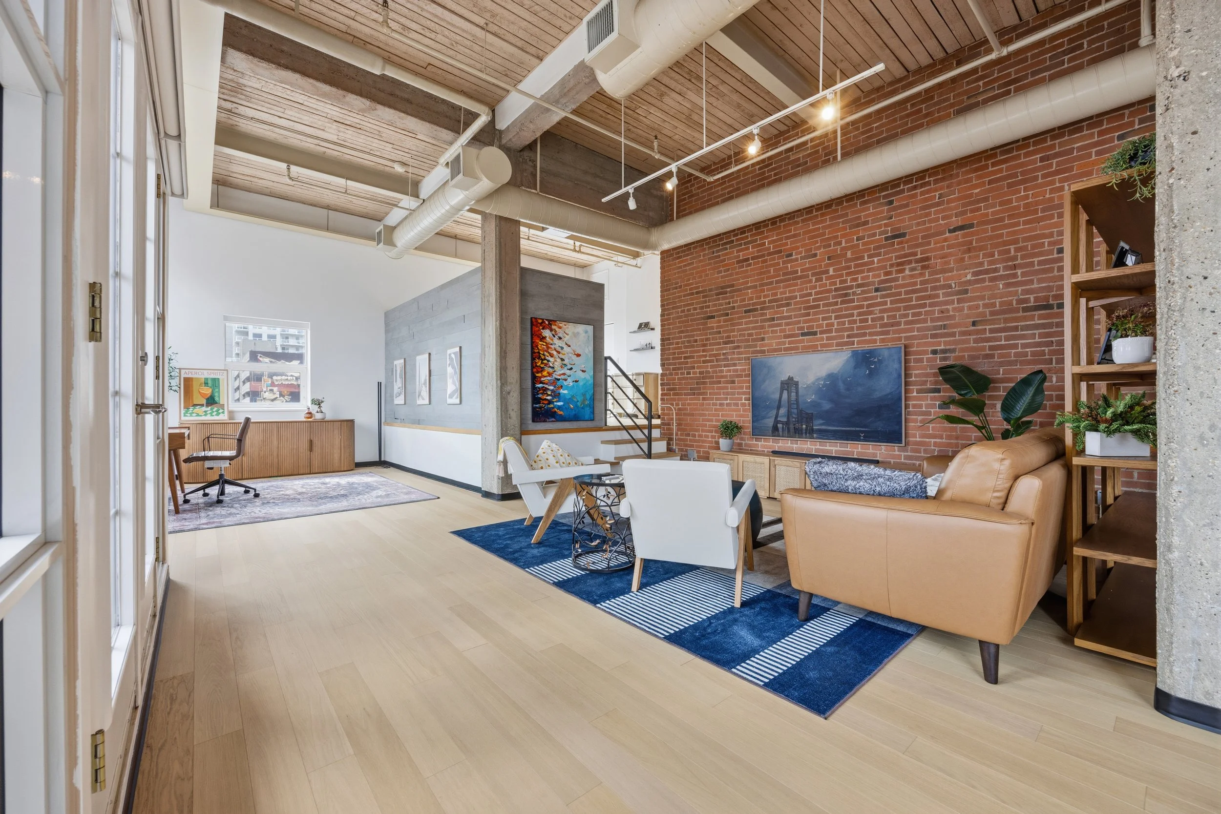 Open-concept living room with exposed brick wall, wooden ceiling, and modern furniture. Features a tan leather sofa, white chairs, and artwork on the walls. There's a large window with a view of a cityscape.