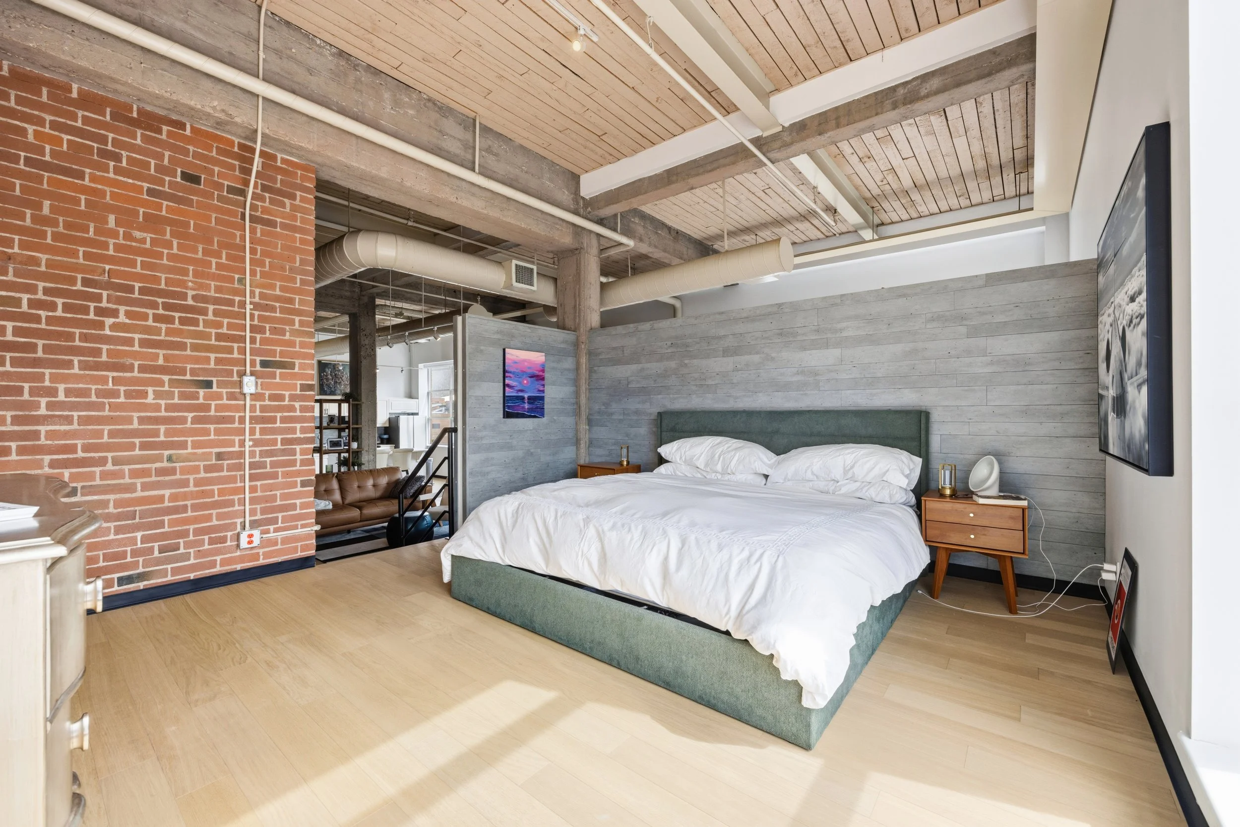 Industrial-style bedroom with a brick wall, wooden ceiling, and ventilated ductwork. A bed with white linens and green bed frame is against a gray wooden wall, with a nightstand and a flat-screen TV mounted on the wall.
