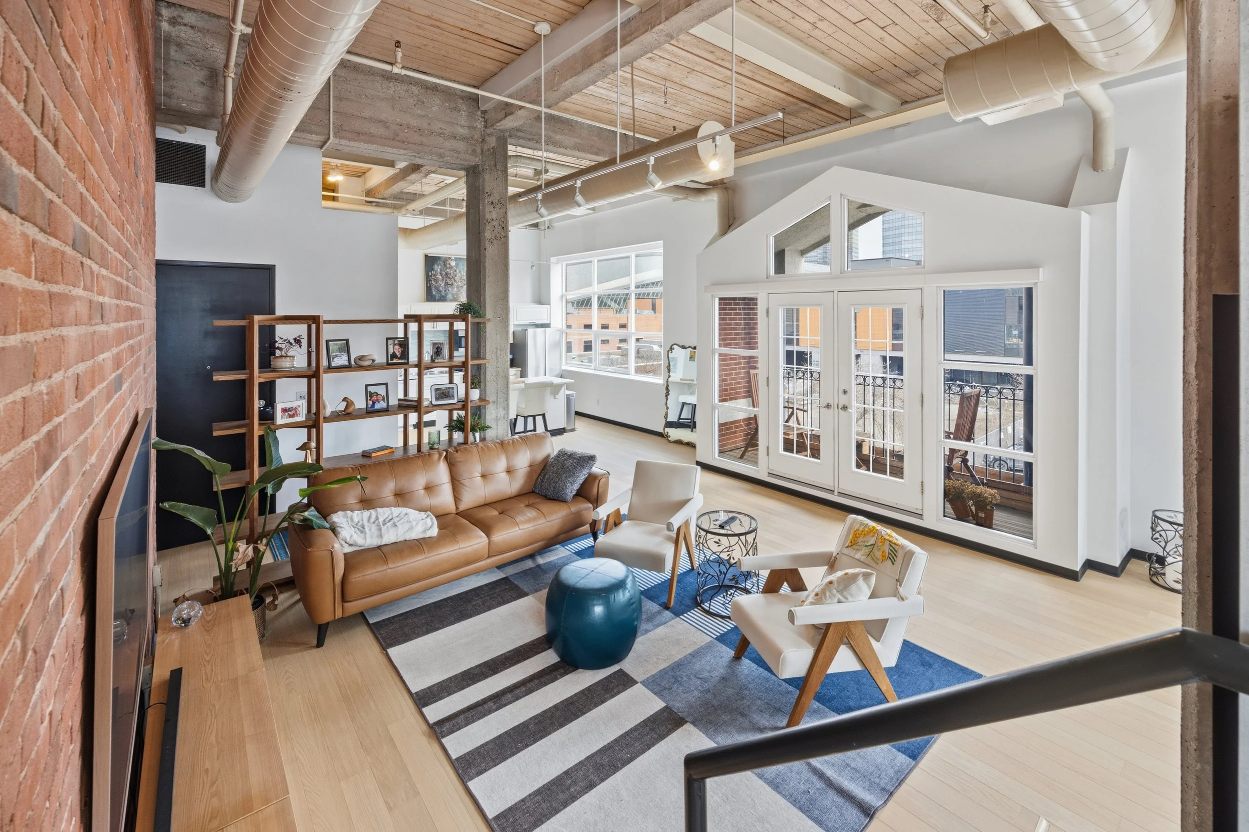 Spacious modern living room with large windows and balcony doors, exposed brick wall, wooden floor, white walls, ceiling with wooden panels and large white air ducts, brown leather sofa, white armchair, geometric area rug, decorative shelving with ph