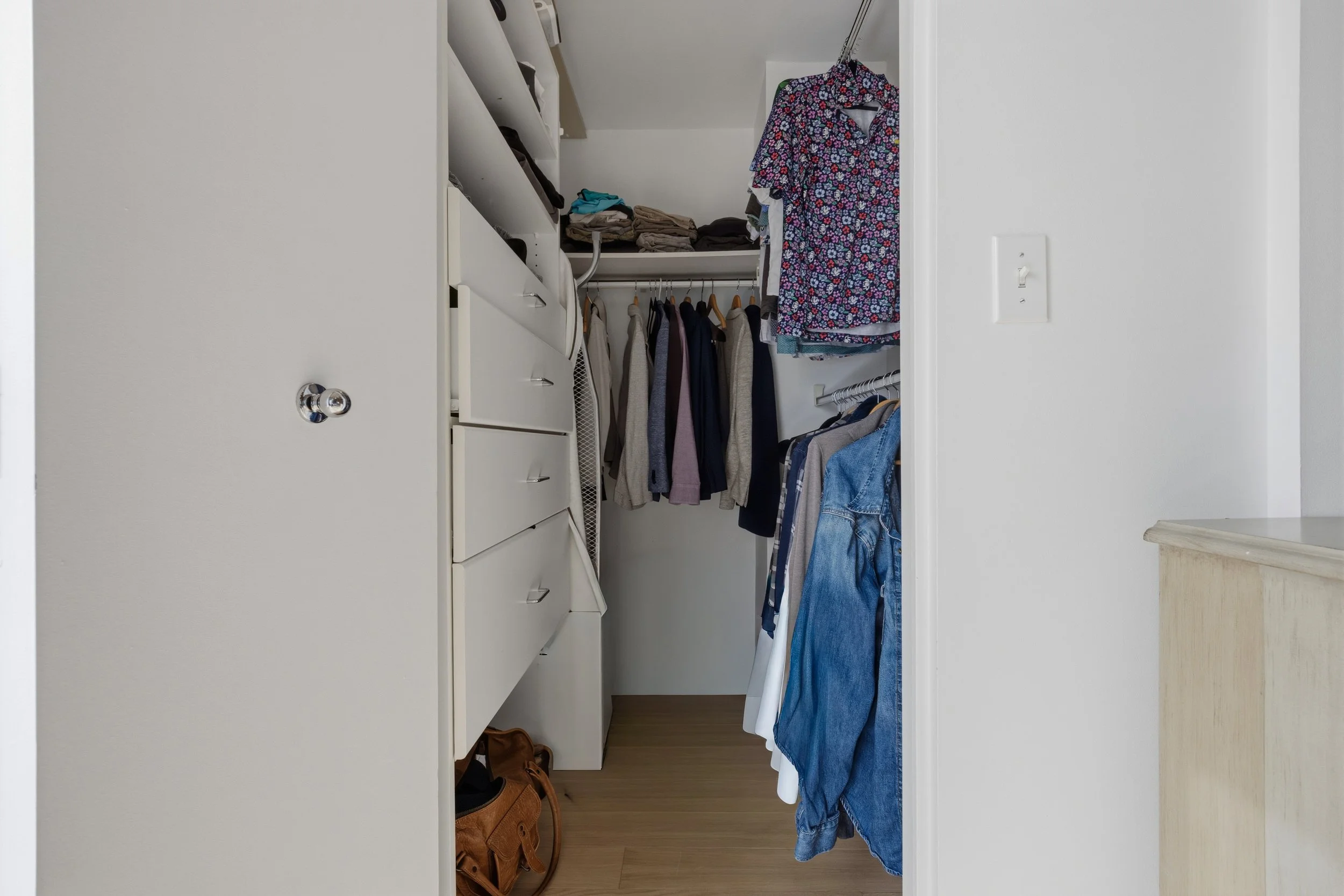 Interior view of a closet with hanging clothes, shelves with folded laundry, and a pair of brown shoes on the floor.