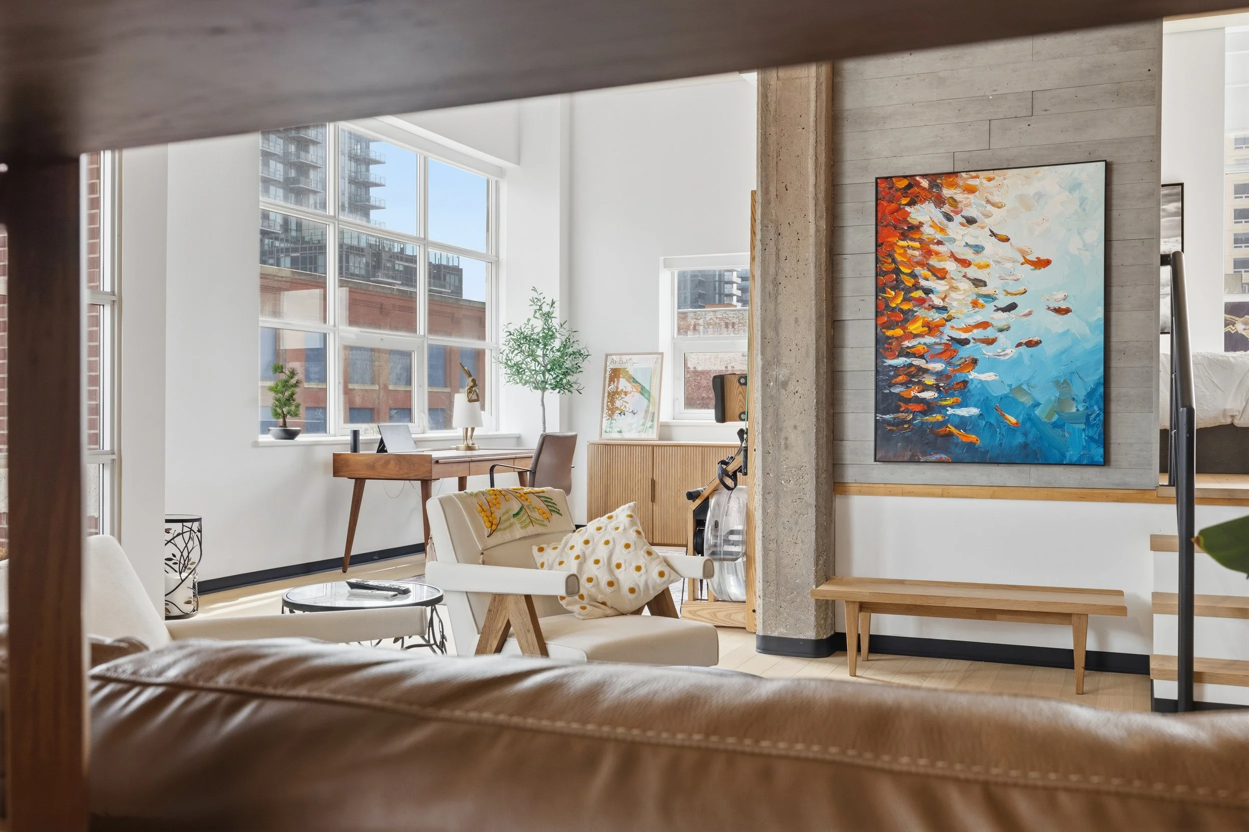 Interior of a modern apartment with large windows, a wooden desk with a lamp, framed artwork, green plants, a white sofa, and a colorful abstract painting on the wall.