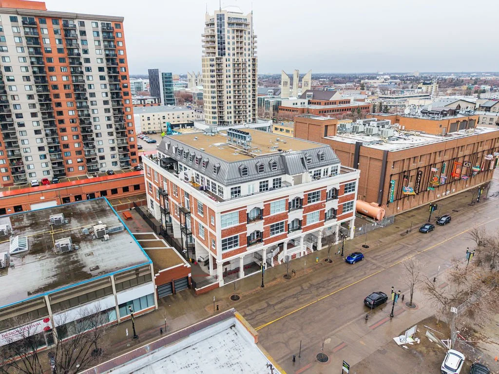 Aerial view of city buildings, including high-rise apartments and a commercial building with colorful signage, on a wet city street with parked cars and streetlights.