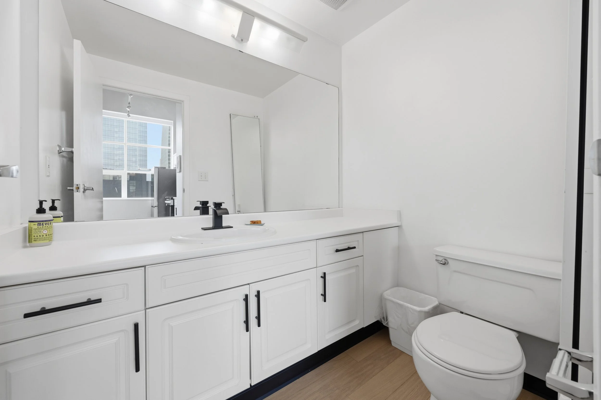 A modern white bathroom with a large mirror, white cabinetry, a white toilet, and a window showing a cityscape outside.