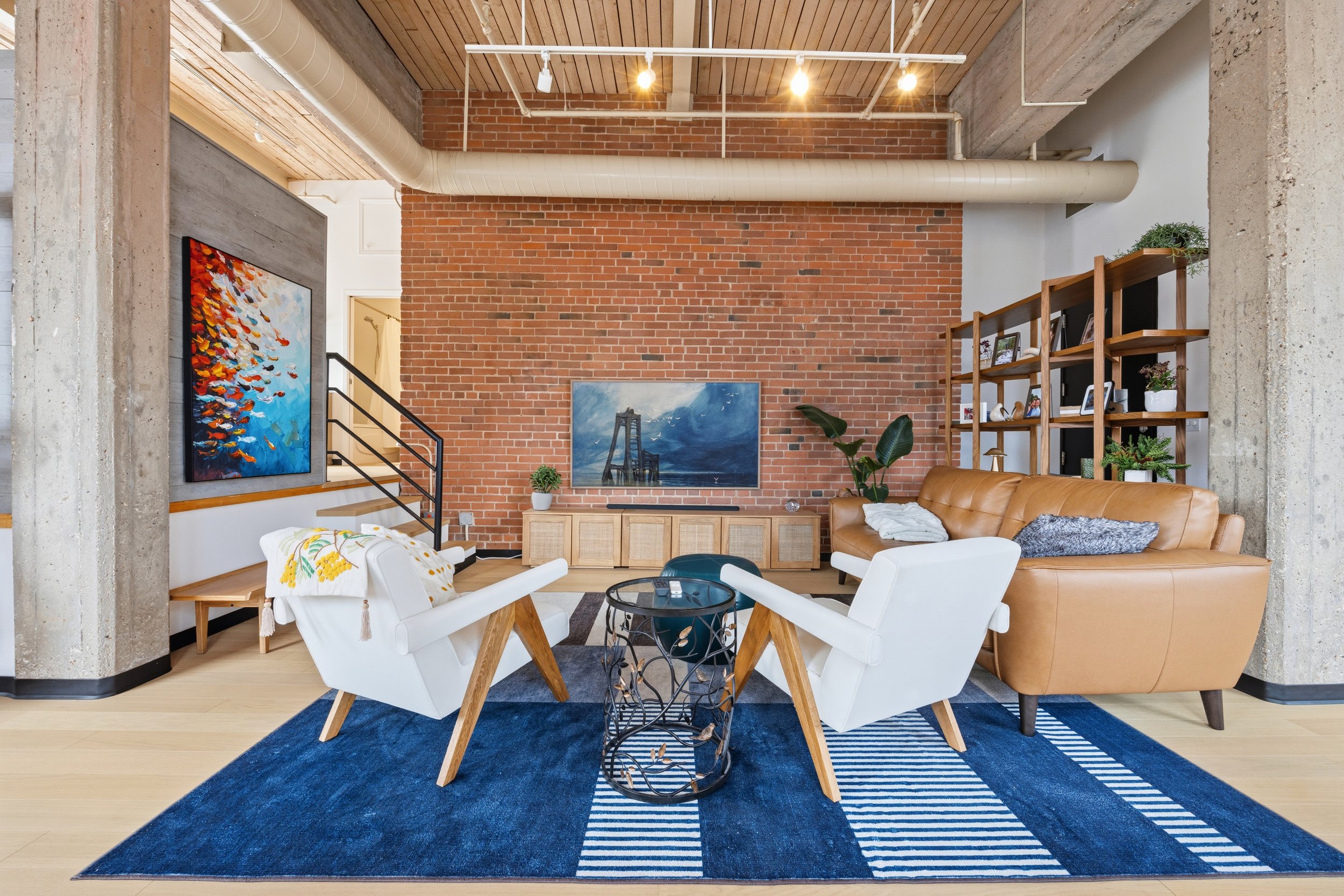 A modern living room with exposed brick walls, a wooden ceiling, and ductwork. It features a leather sofa, white armchairs, artwork on the walls, and a blue patterned rug.