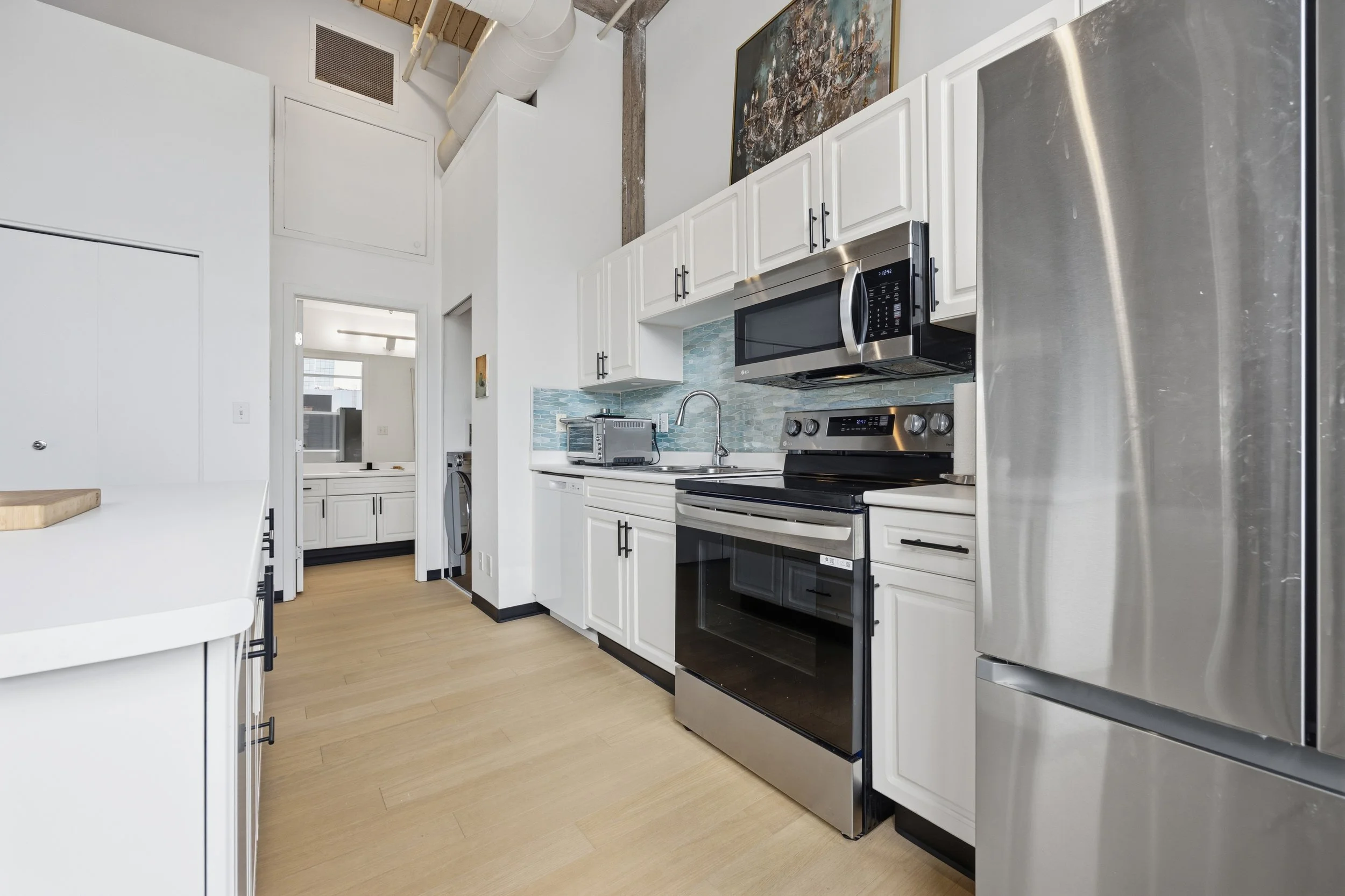 Modern kitchen with white cabinets, stainless steel refrigerator, microwave, oven, and light wood flooring.