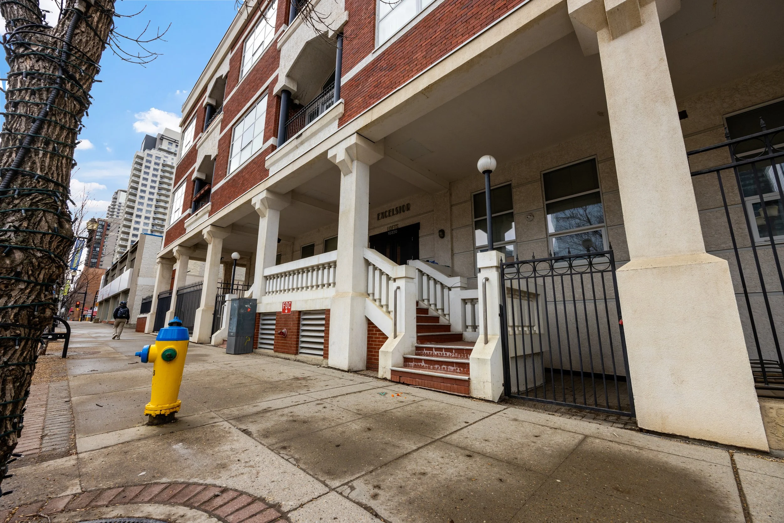 Street view of a multi-story apartment building with red brick and beige concrete accents, a staircase leading to the entrance, a yellow fire hydrant on the sidewalk, tall trees, and high-rise buildings in the background.