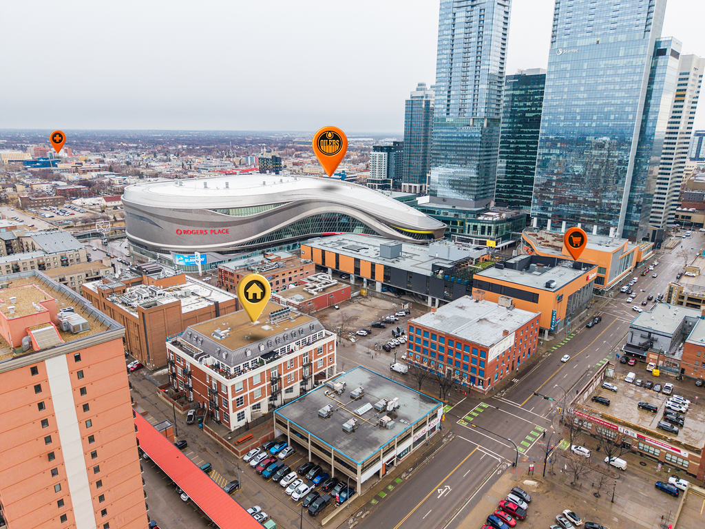 A cityscape view featuring Rogers Place arena, a large sports and entertainment complex, with several modern high-rise buildings and parking lots around it. The scene shows an urban environment with streets, cars, and various buildings.