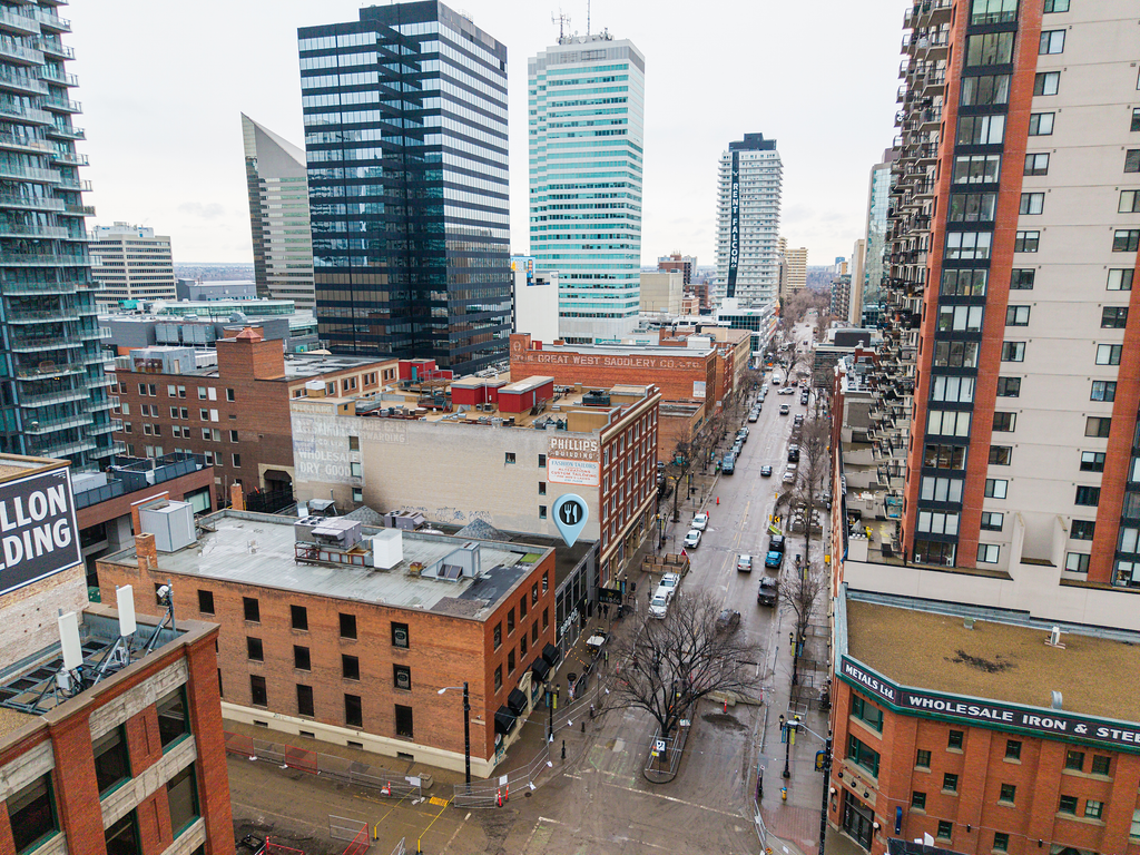 A city landscape with high-rise buildings, city streets with parked and moving cars, and some leafless trees on a cloudy day.