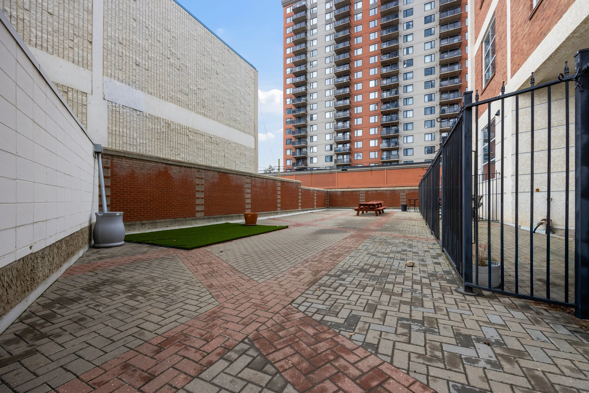 An urban courtyard with brick and concrete pavement, a small patch of artificial grass, a picnic table, and a black metal fence. Surrounding buildings include a large beige brick structure, a white wall, and a tall apartment building with balconies u