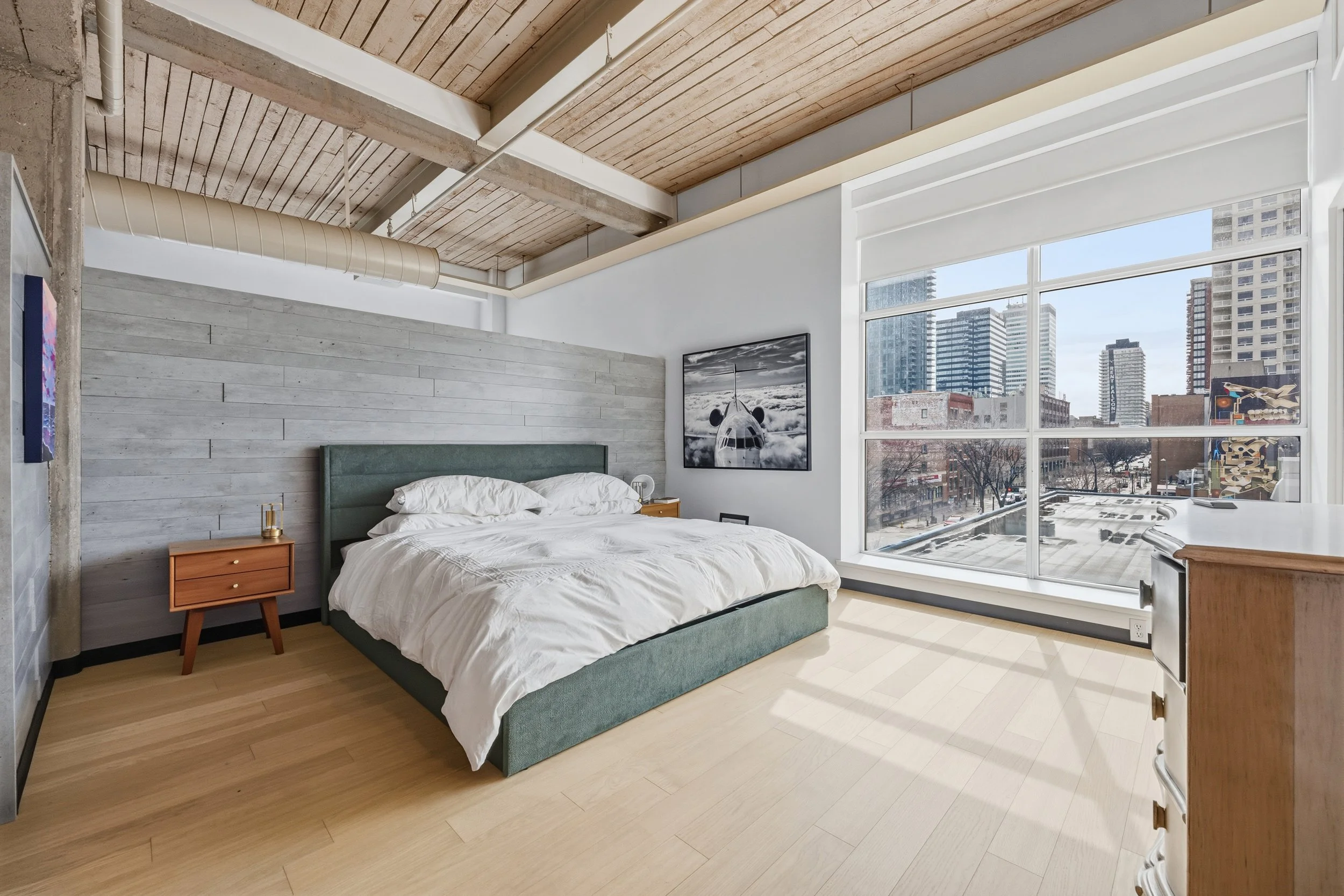 Modern bedroom with large window showing cityscape, bed with white bedding, wooden nightstand, and art on walls.