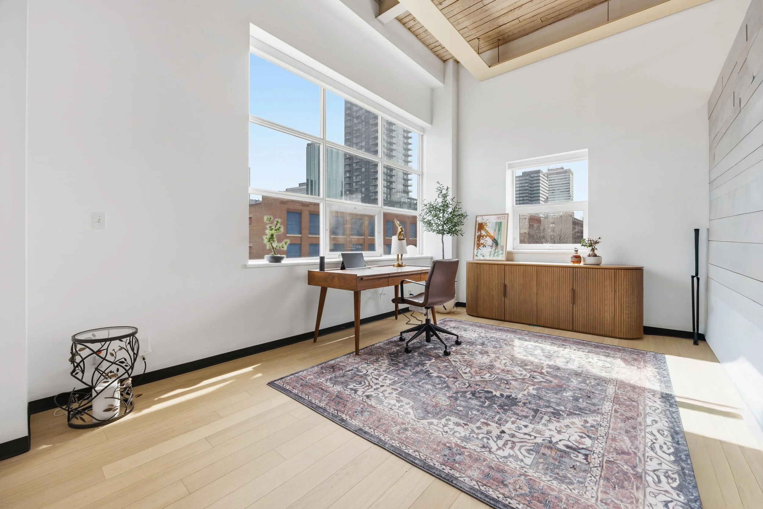 Bright office with large windows showing city buildings, wooden desk with chair, plants, artwork, and a patterned rug.