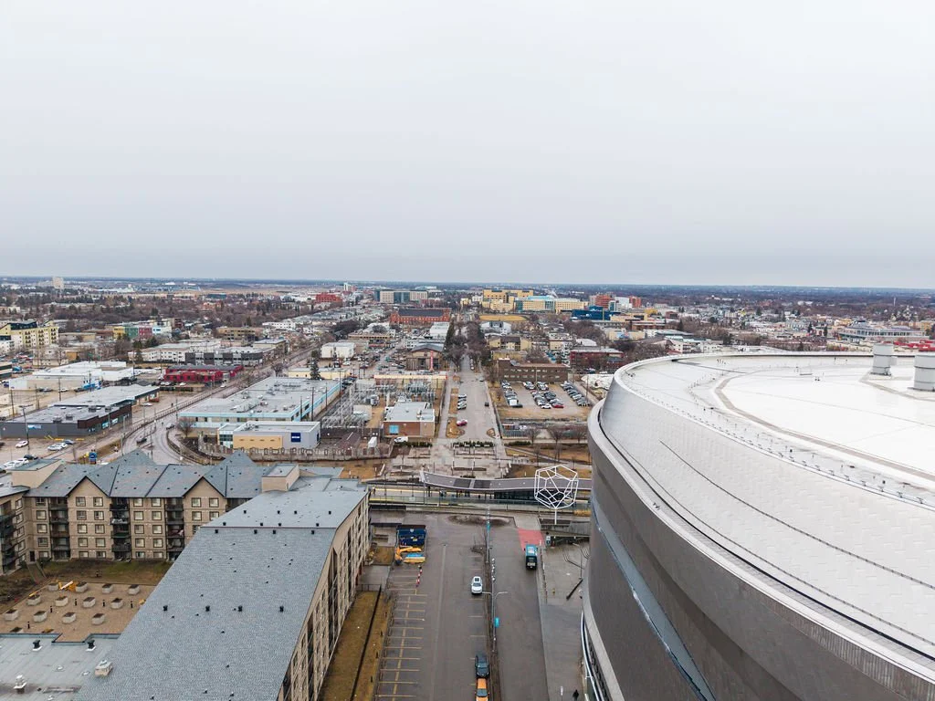A cityscape viewed from a high-rise building, showing a parking lot, streets, and buildings, with a curved rooftop in the foreground and an overcast sky.