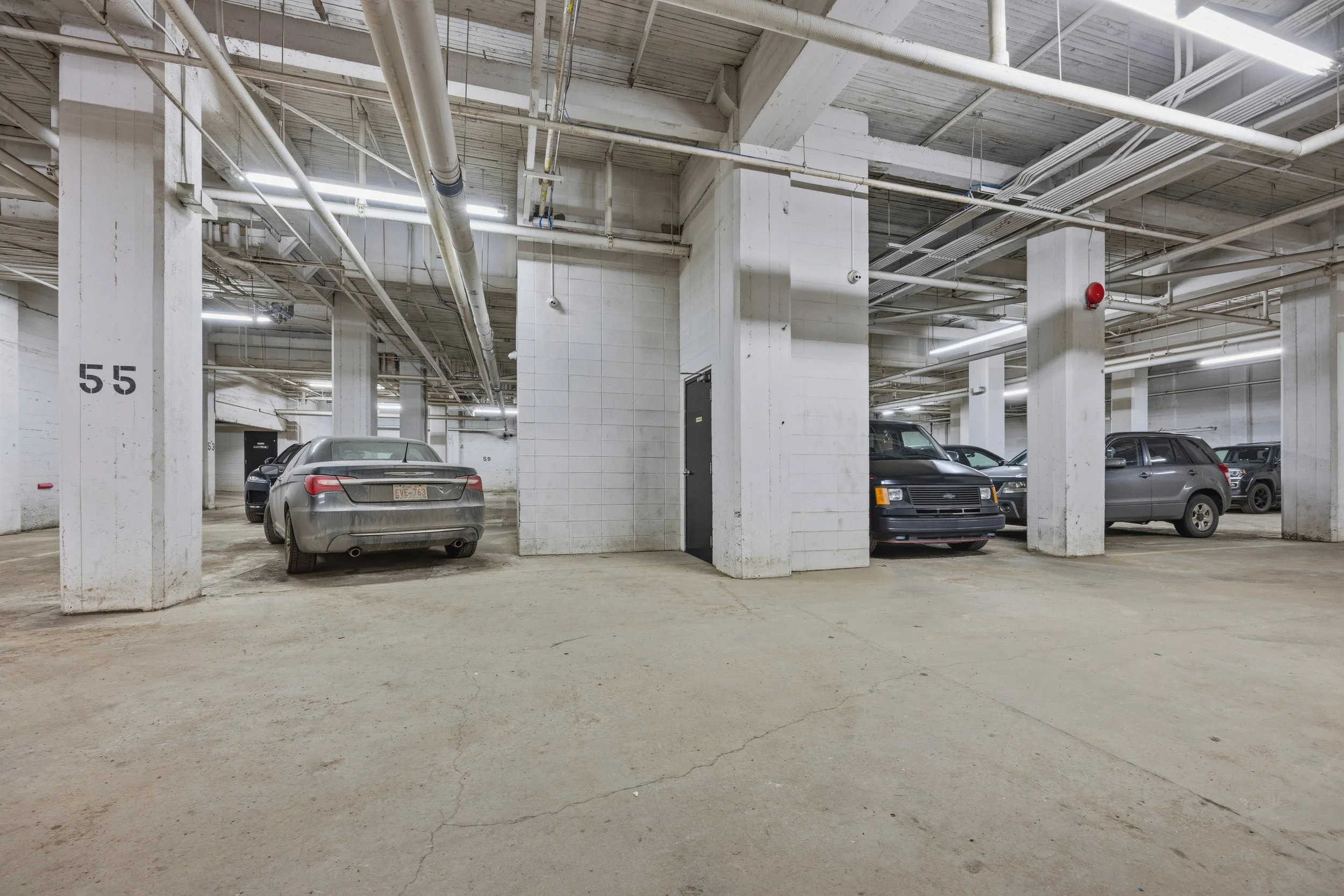 Underground parking garage with concrete pillars and several parked cars, including sedans and an SUV. The ceiling has exposed pipes and fluorescent lighting.