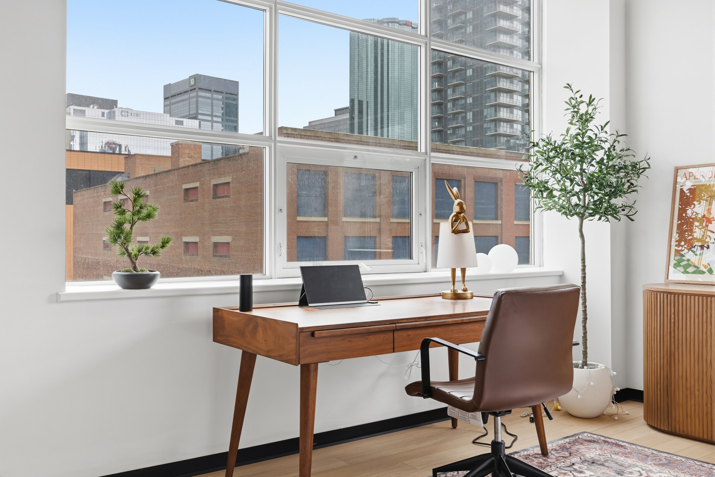 Modern office with large windows, wooden desk and chair, potted plants, decorative items, and cityscape view.
