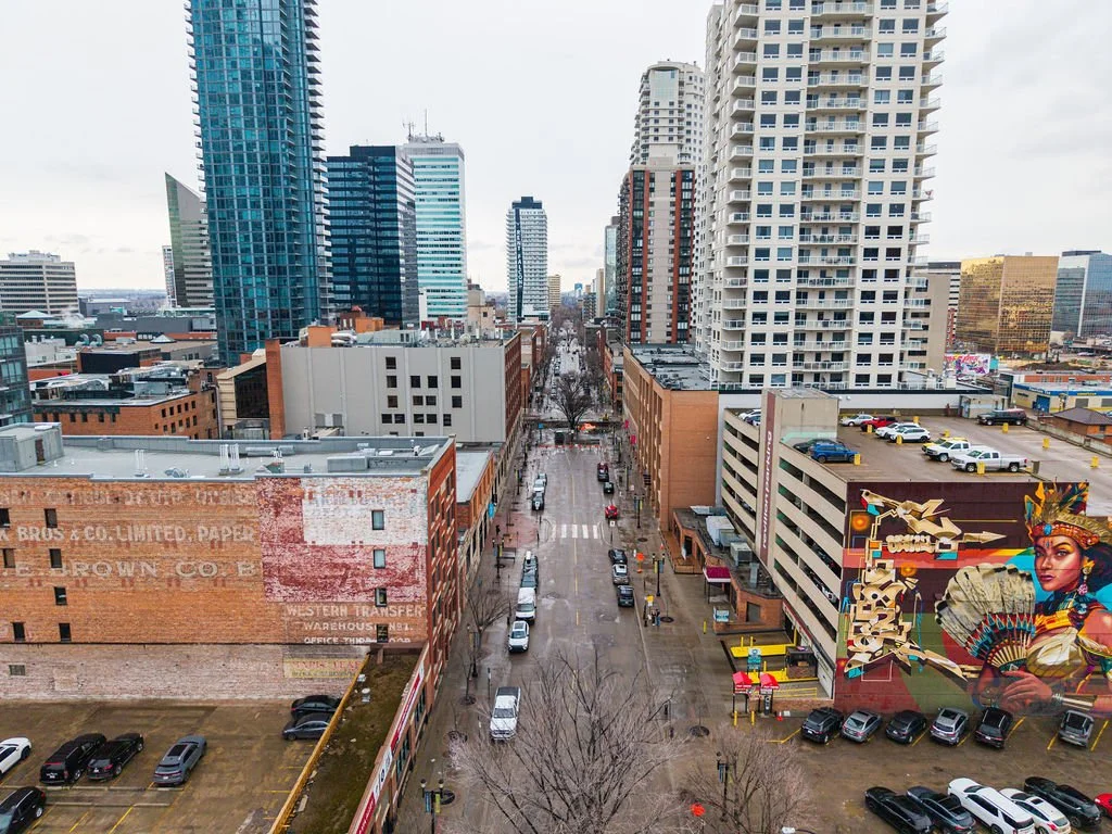City street view with tall modern buildings, a mural of a woman in traditional attire, and parked cars.