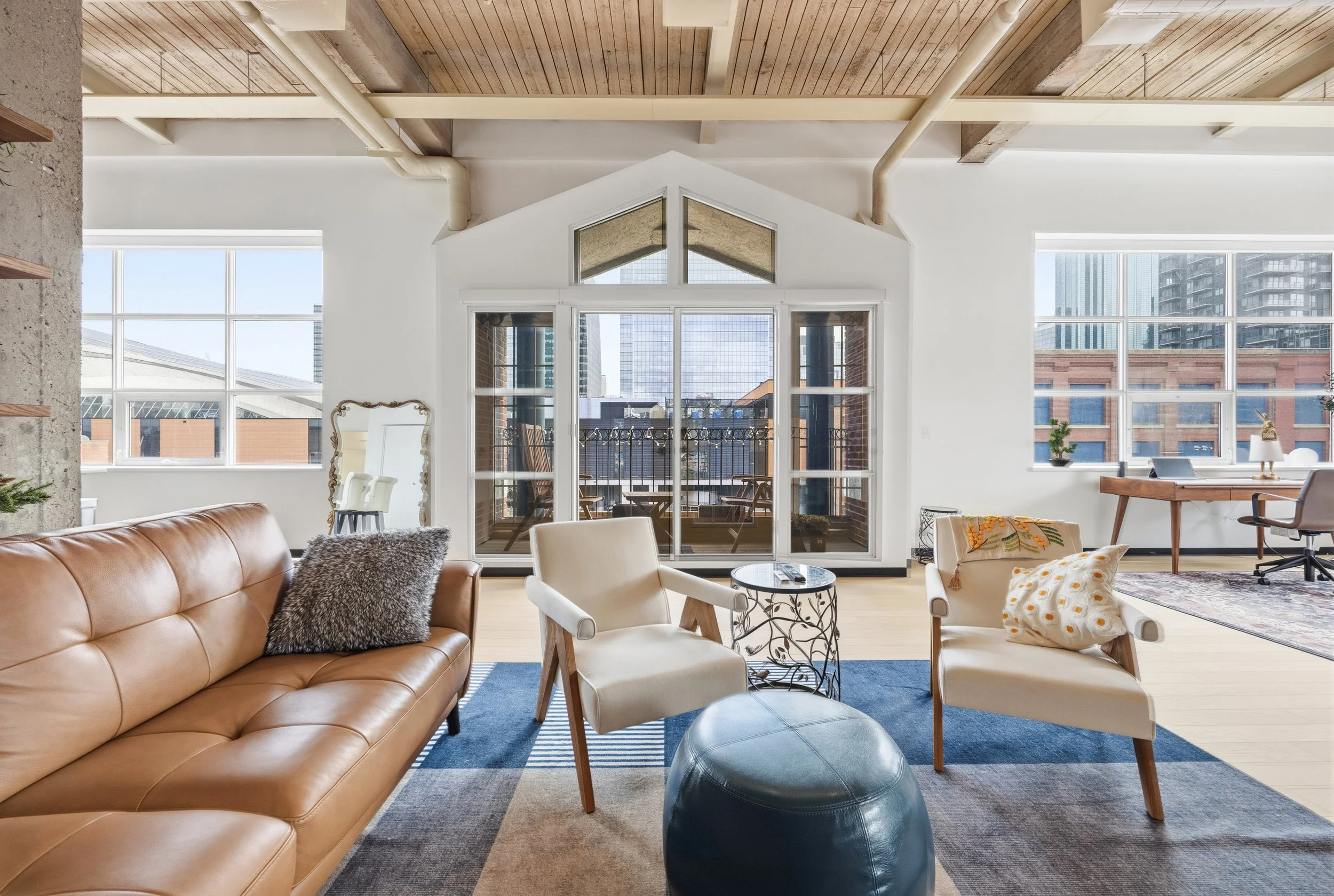 Spacious modern living room with large windows showing city view, featuring leather sofa, armchairs, rug, and small table.