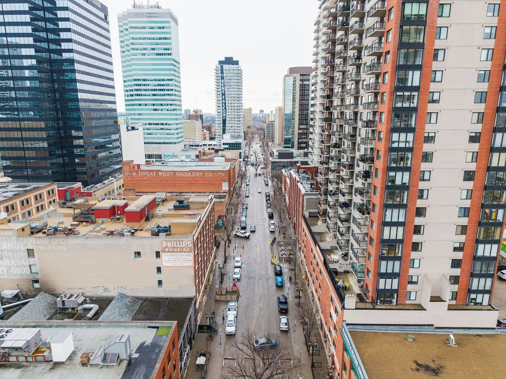A city street view with tall office buildings and apartment complexes, parked cars along the sides, and leafless trees, under an overcast sky.