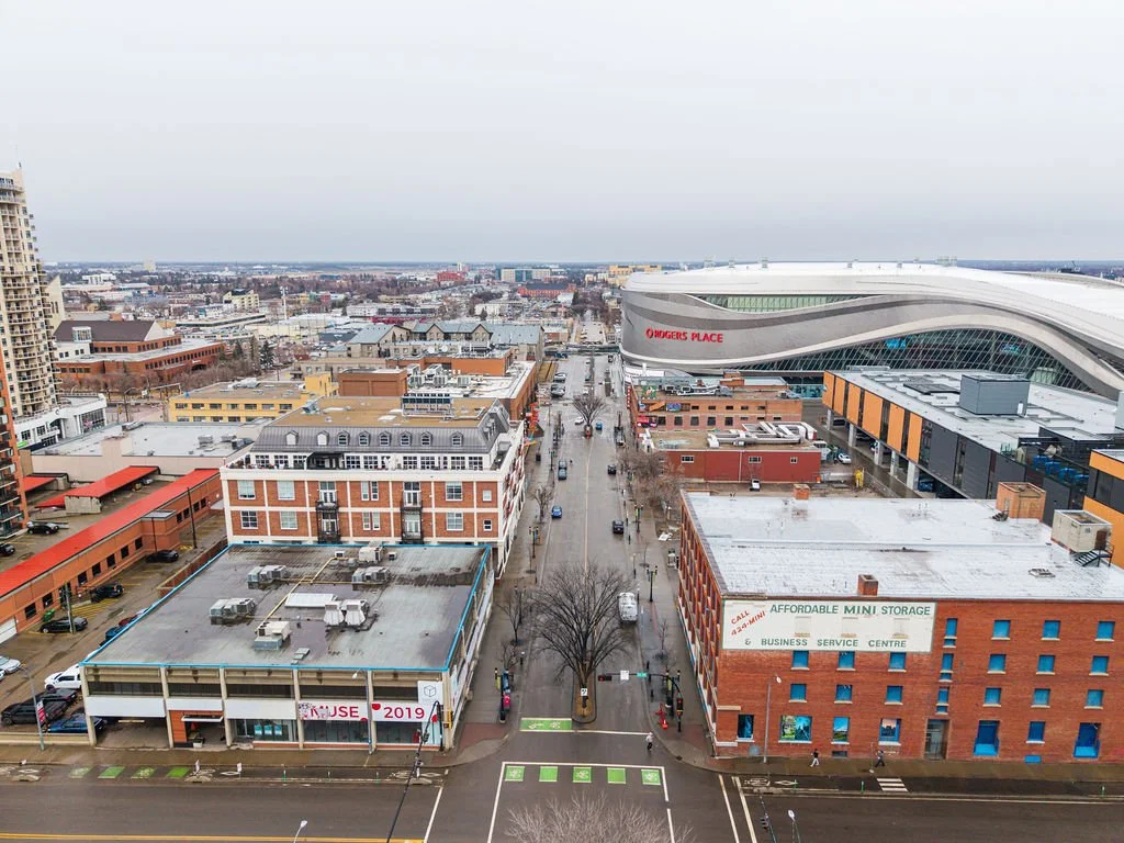 City street view featuring buildings, a large modern sports arena labeled 'Rogers Place,' and parking lots, taken on a cloudy day.