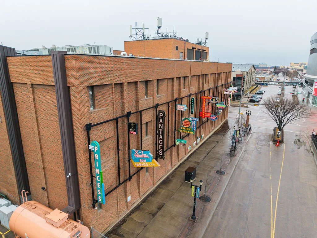A brick building with multiple colorful signs hanging on the exterior, including signs for a pawn shop, printers, and various other businesses, on a rainy day in an urban area.