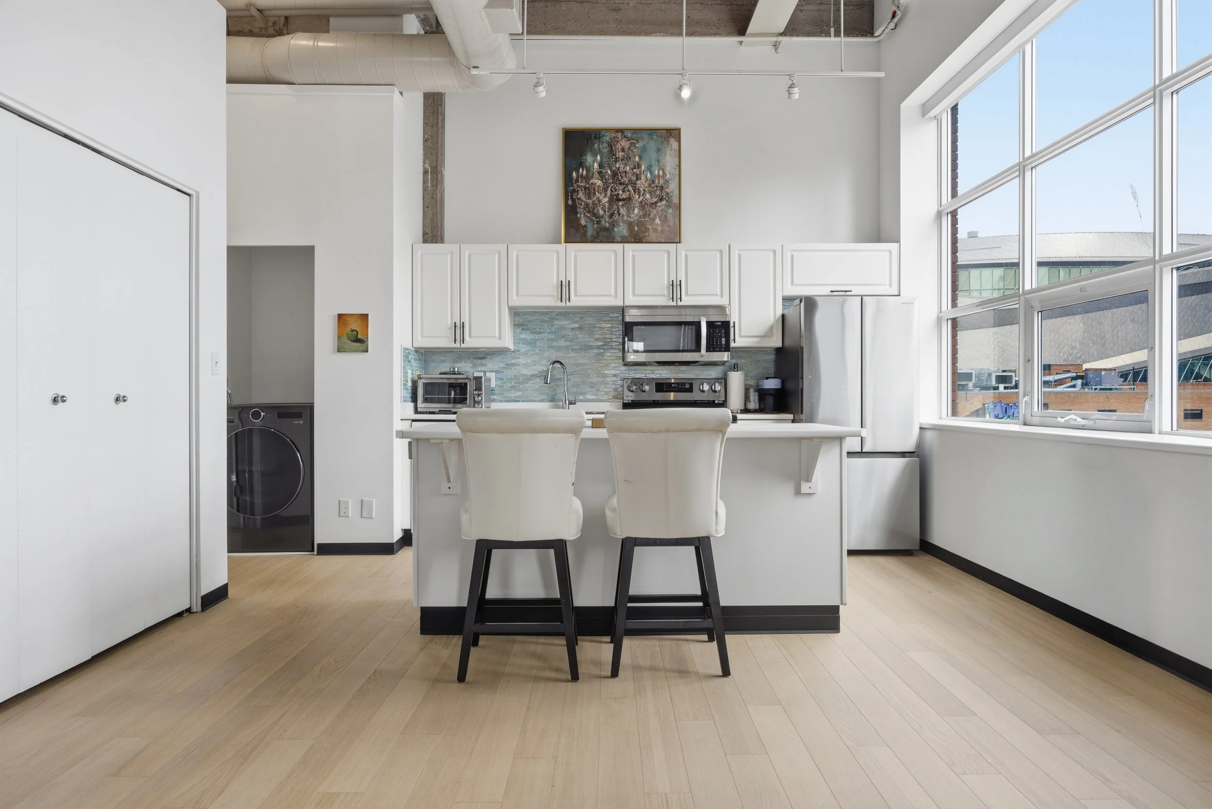 Modern kitchen with white cabinets, light wood flooring, and large windows providing natural light. Contains a microwave, oven, and small appliances on the counter, with two white chairs at a white island.