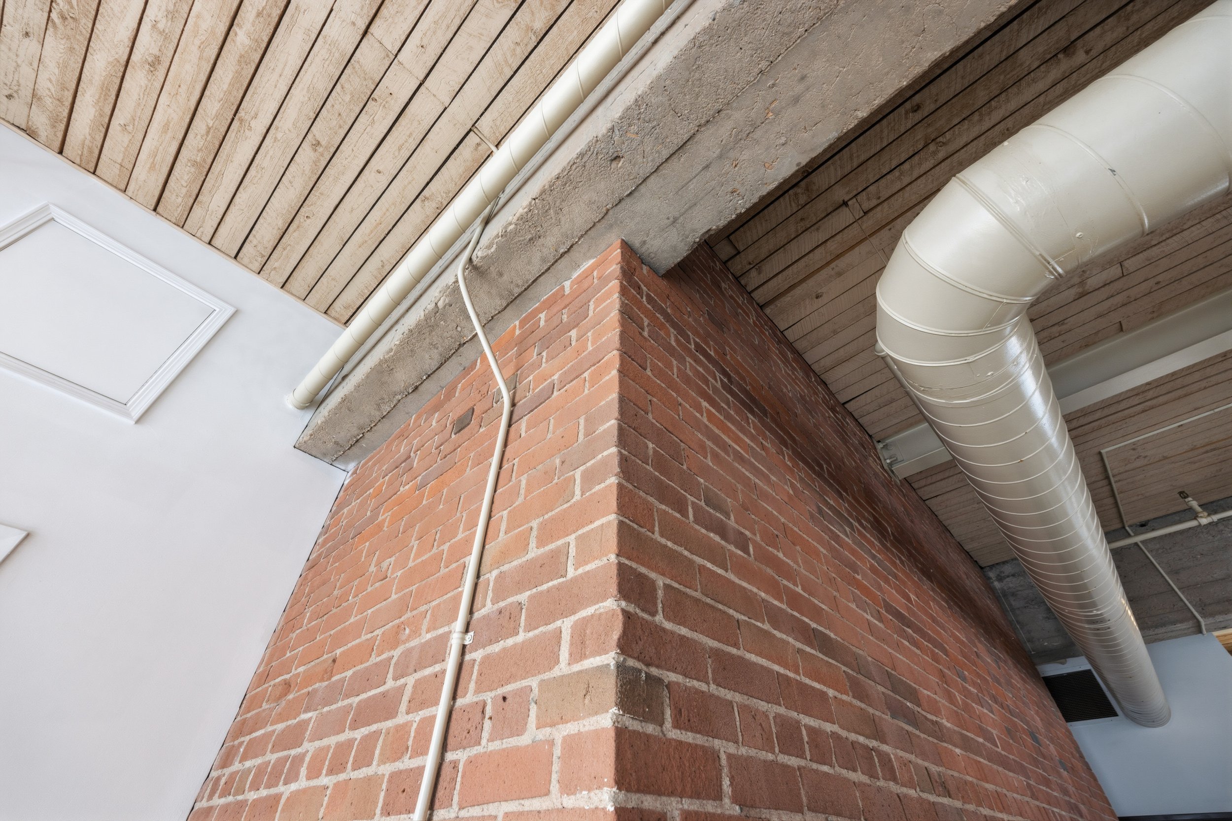 Close-up of building corner showing brick wall, concrete beam, wooden ceiling, and white pipes.