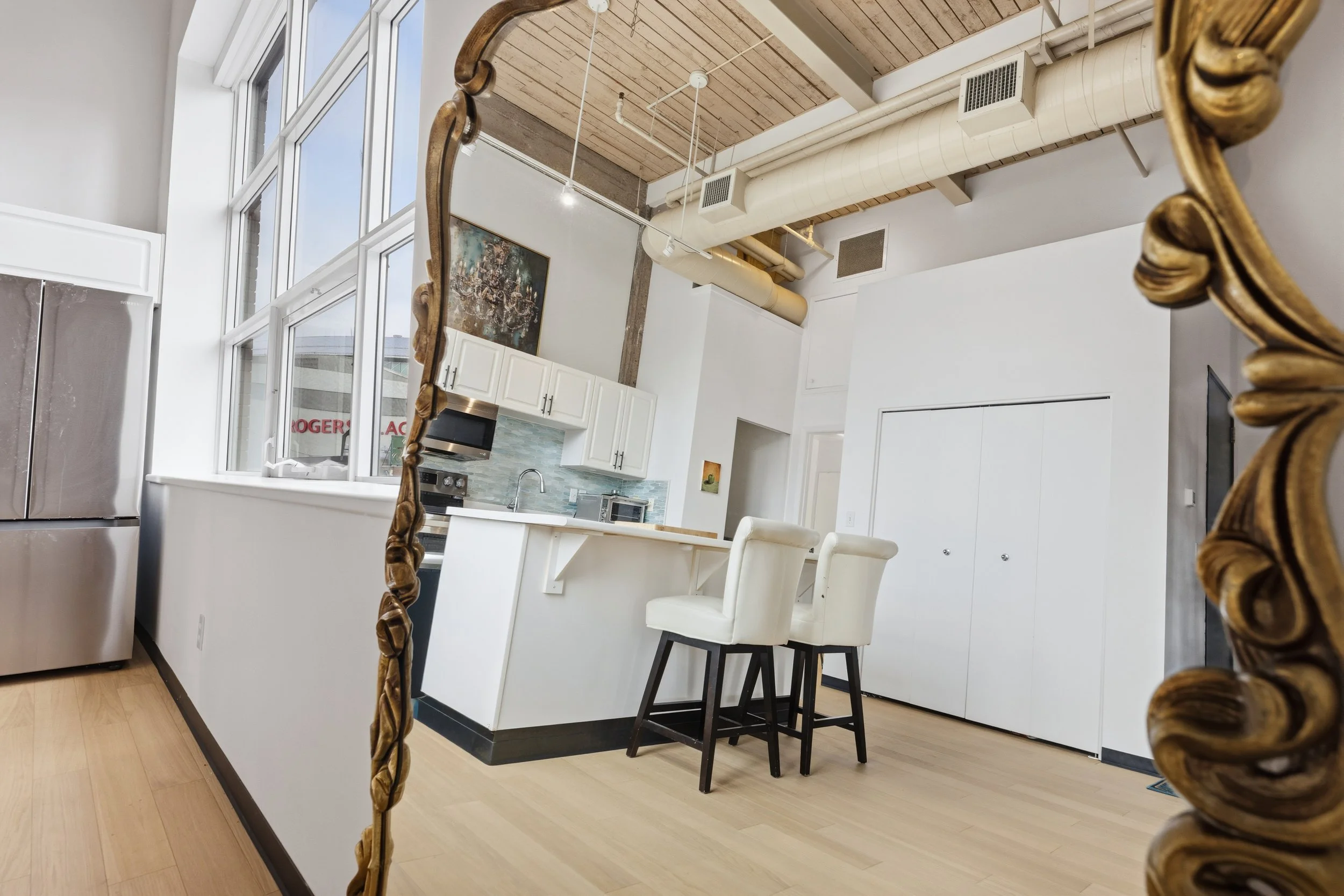 Interior of a modern kitchen with white cabinets, a small breakfast bar with two white chairs, a stainless steel refrigerator, and large windows allowing natural light, with an exposed ceiling and ductwork.
